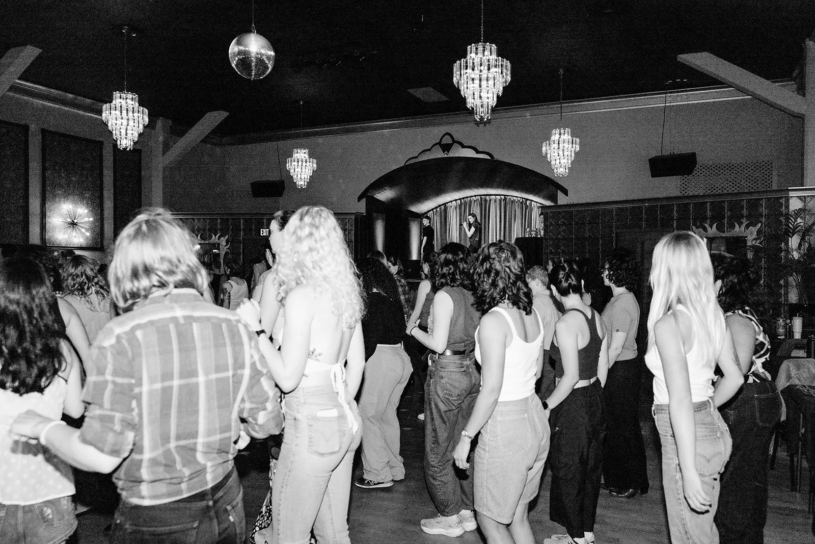 Group of people standing and facing a stage with performers in a nightclub or event space, with chandeliers hanging from the ceiling.