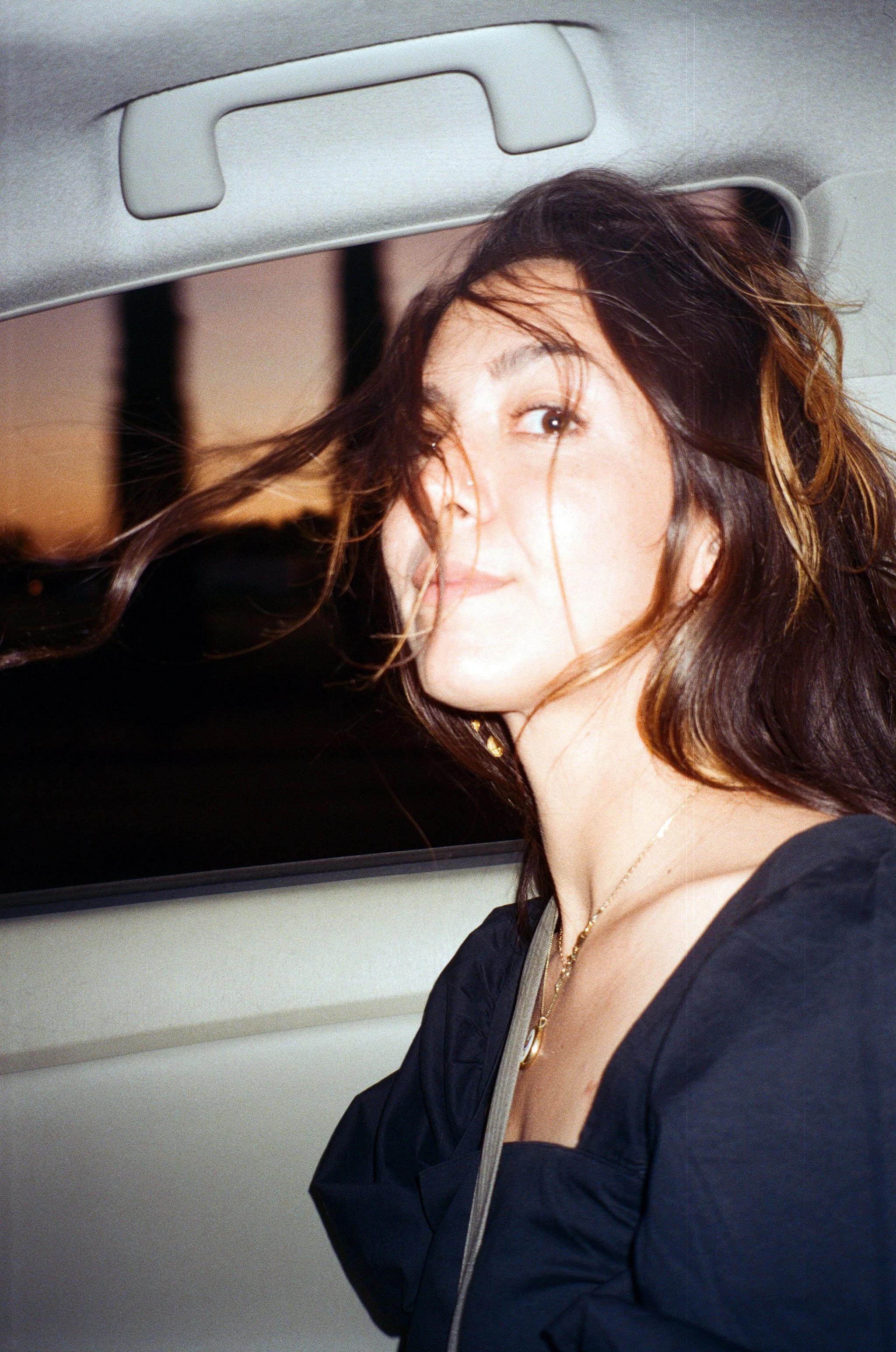 A woman with dark brown hair and a black top sitting in a vehicle at sunset, looking at the camera with windblown hair.