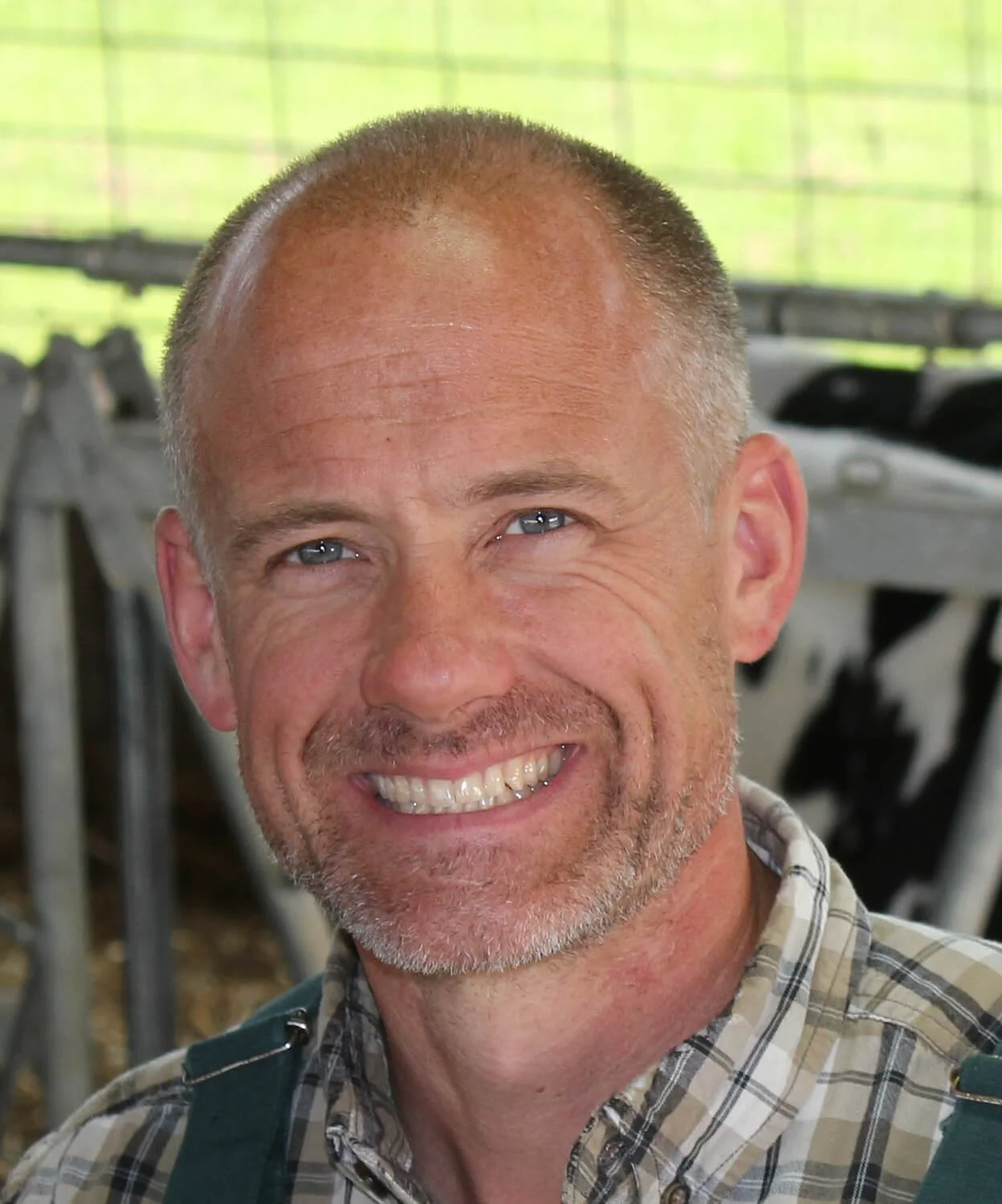 A smiling middle-aged man with short gray hair and blue eyes, wearing a plaid shirt, standing inside a barn with blurred farm equipment in the background.