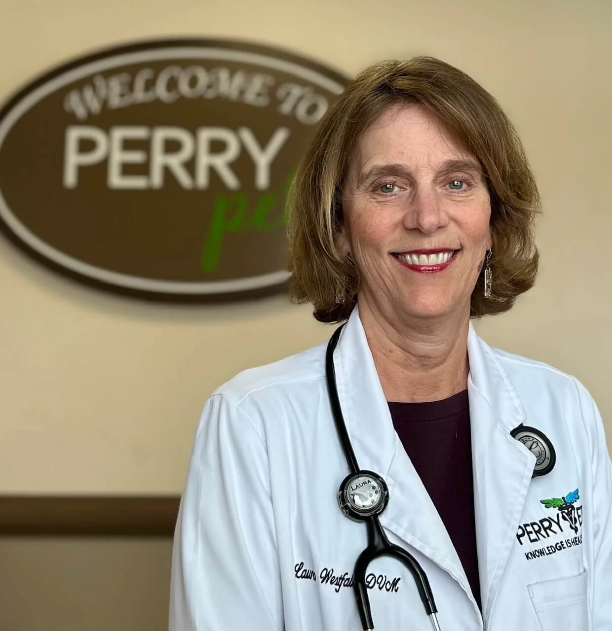 A smiling woman in a white medical coat with a stethoscope around her neck, standing in front of a sign that reads 'Welcome to Perry' in a medical office or clinic.