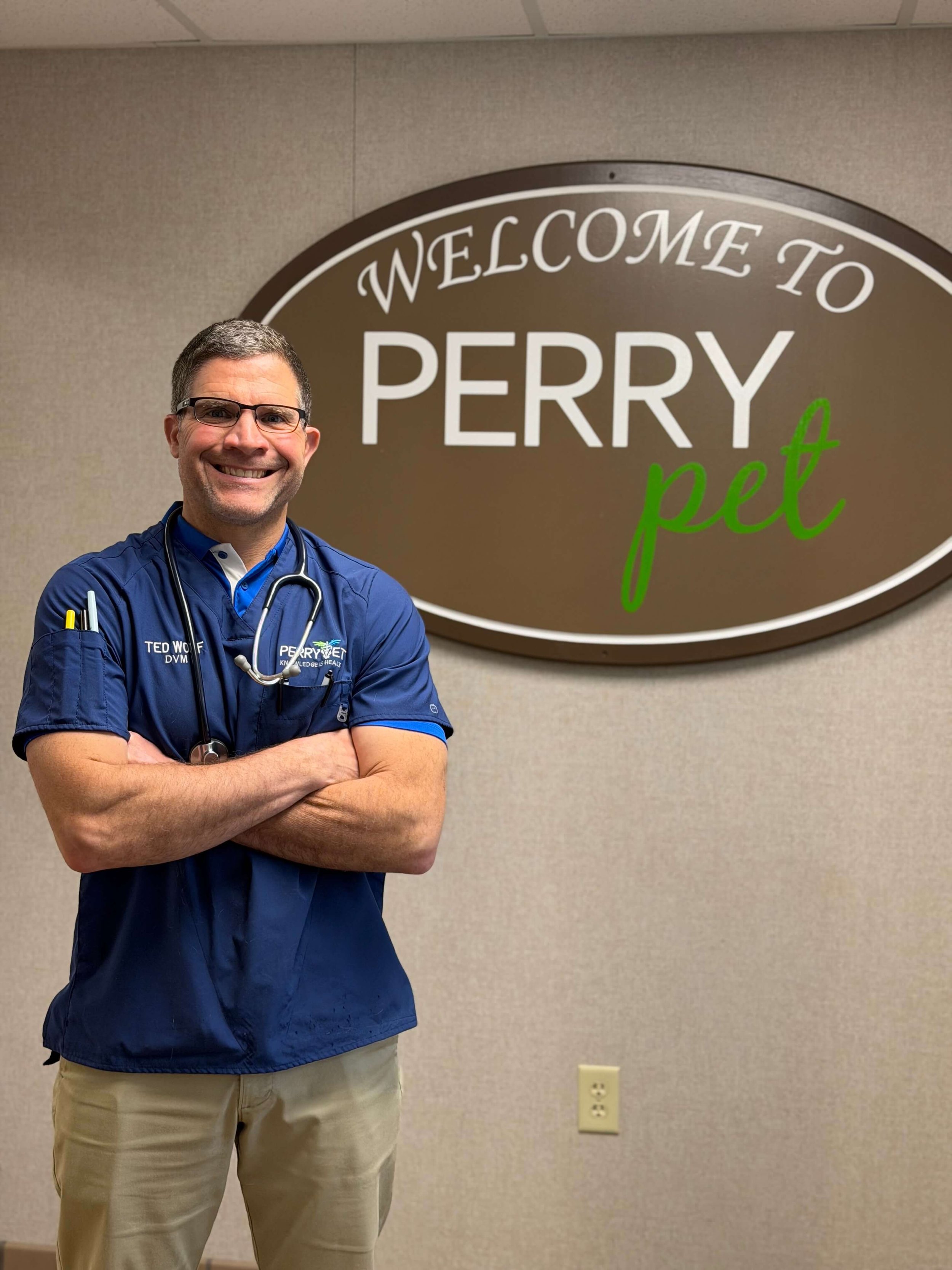A man wearing glasses and a blue veterinary uniform with a stethoscope around his neck, smiling and standing with arms crossed in front of a sign that reads 'Welcome to Perry Pet' on a beige wall.