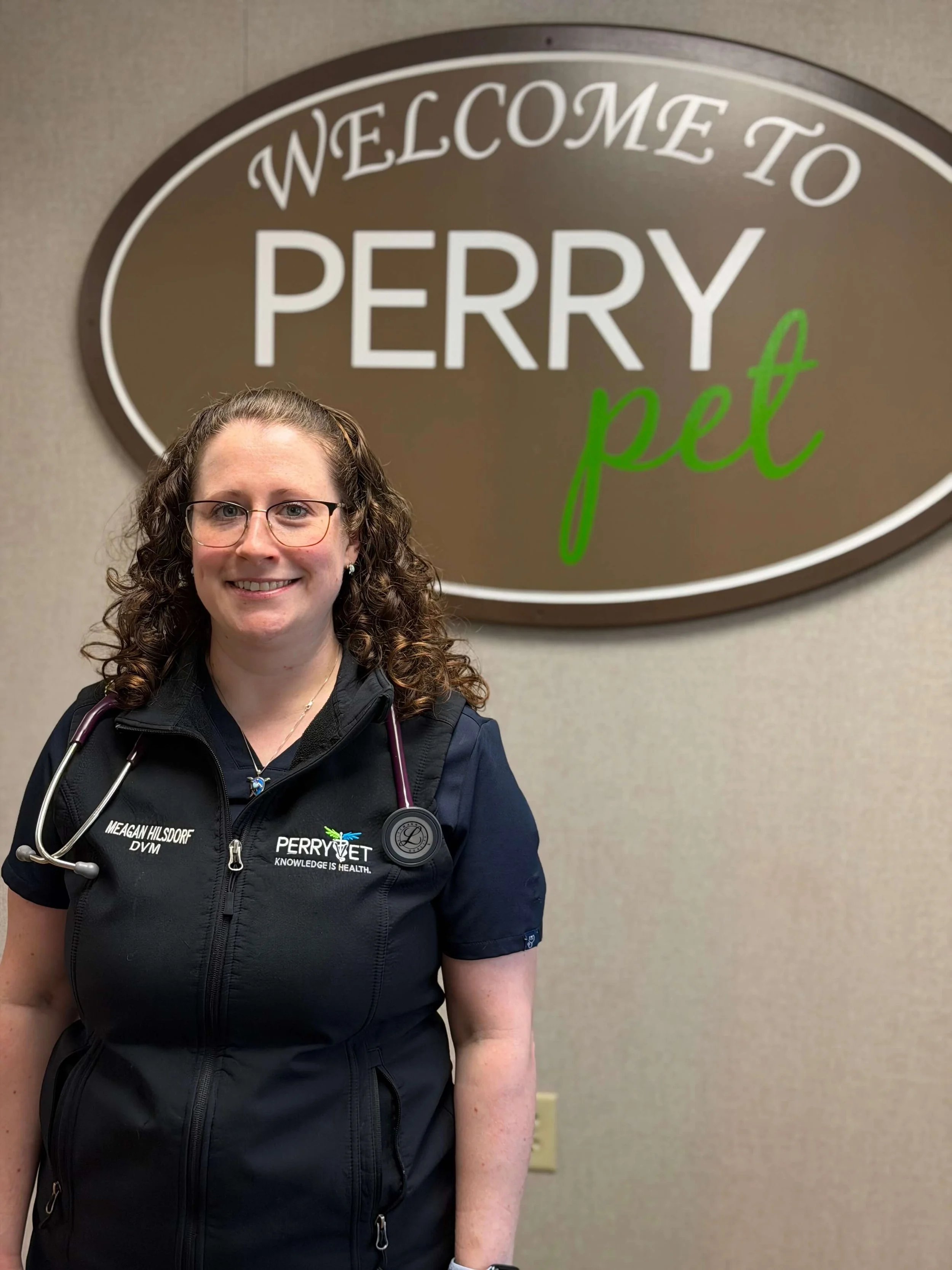 A woman with curly hair, glasses, and a stethoscope around her neck standing in front of a sign that reads "Welcome to Perry Pet" on a beige wall.