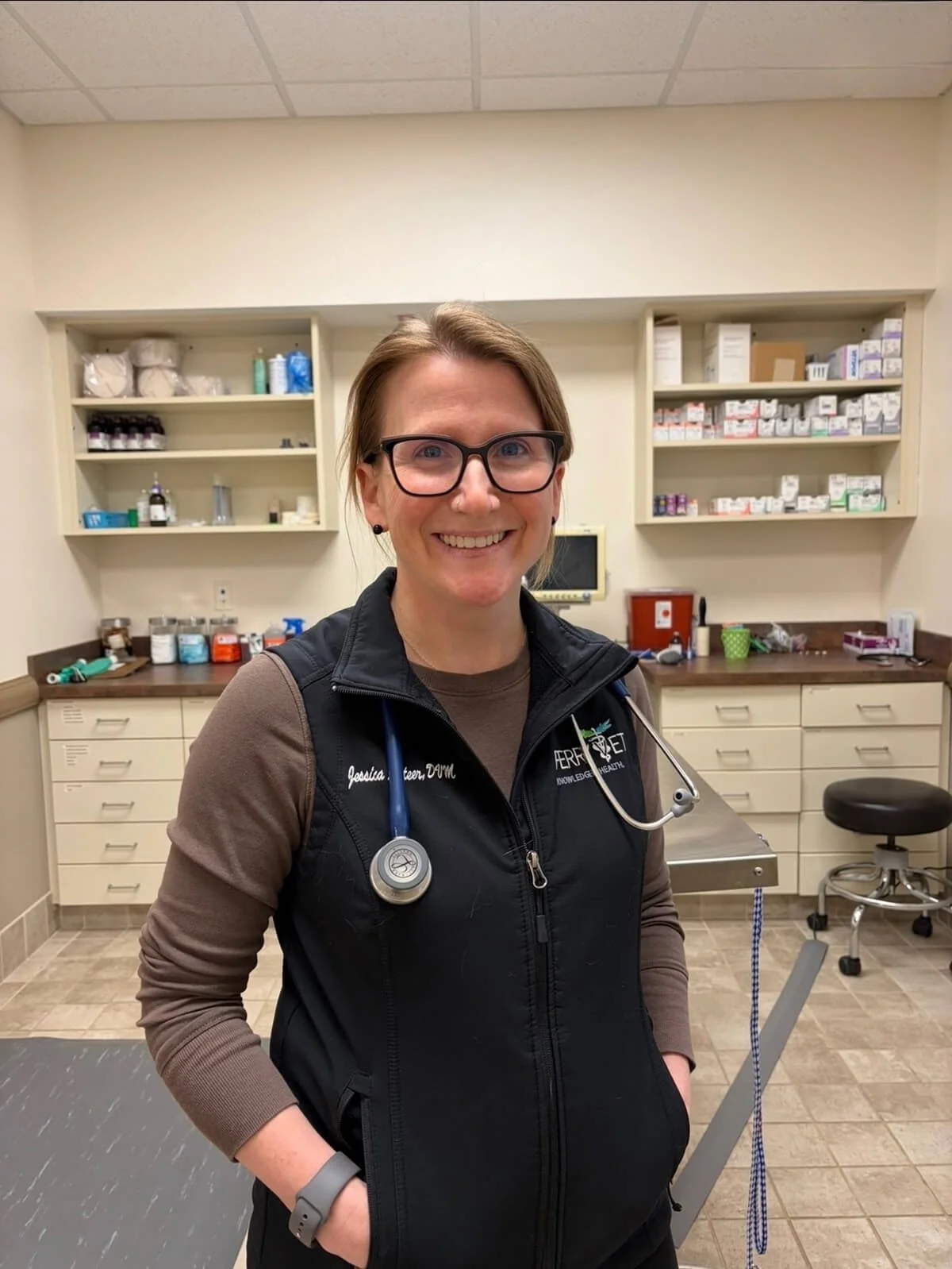 A smiling woman wearing glasses and a black vest with a stethoscope around her neck, standing in a medical office with medical supplies and equipment in the background.