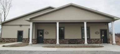 A small, single-story building with a front porch, white pillars, and stone accents at the bottom of the walls, under a sloped roof.