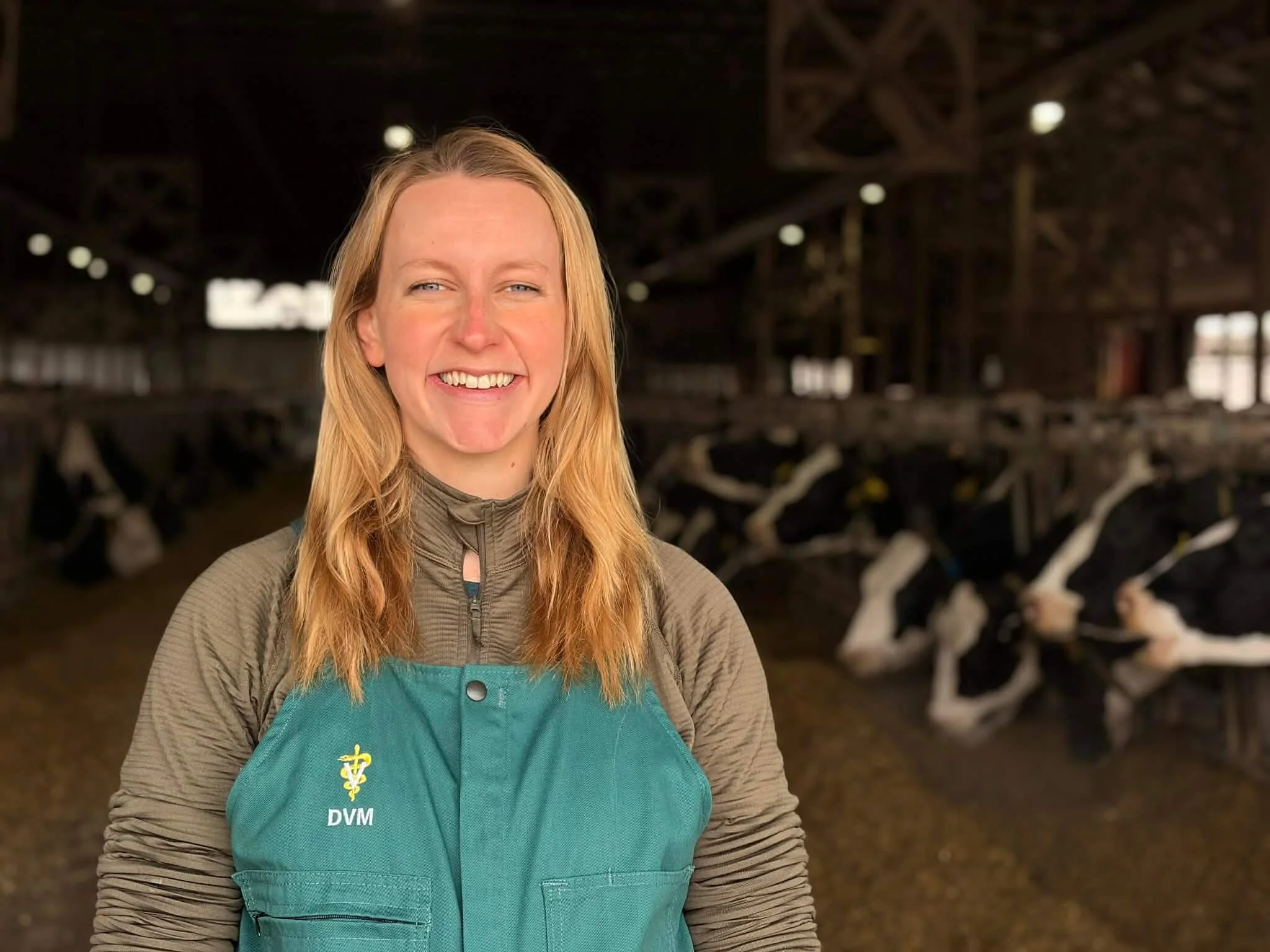 A woman with blonde hair smiling, wearing a teal veterinary coat with a medical symbol and the letters DVM, standing inside a barn with cows in the background.