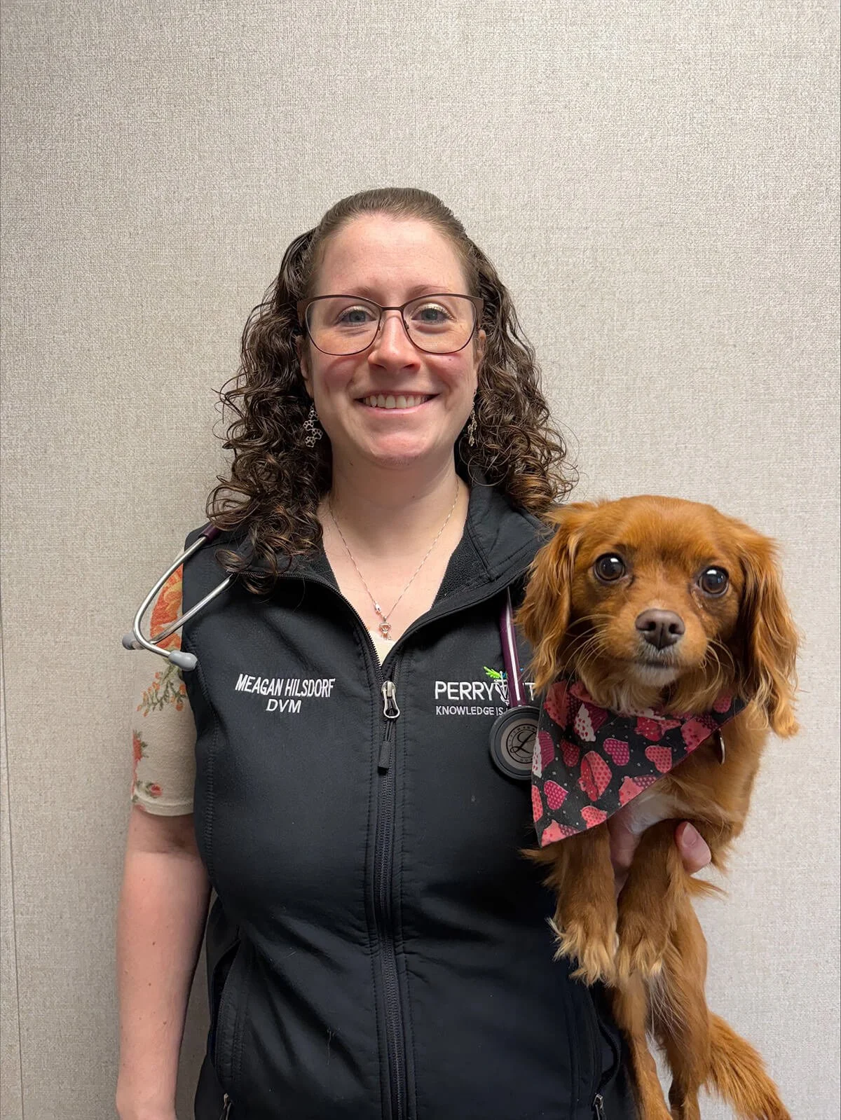 A woman with curly hair, glasses, and a stethoscope around her neck standing in front of a sign that reads "Welcome to Perry Pet" on a beige wall.