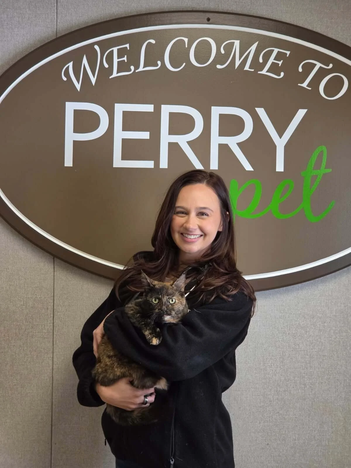 Woman with long brown hair smiling and holding a tortoiseshell cat in front of a large sign that says "Welcome to Perry Pet" on a gray wall.