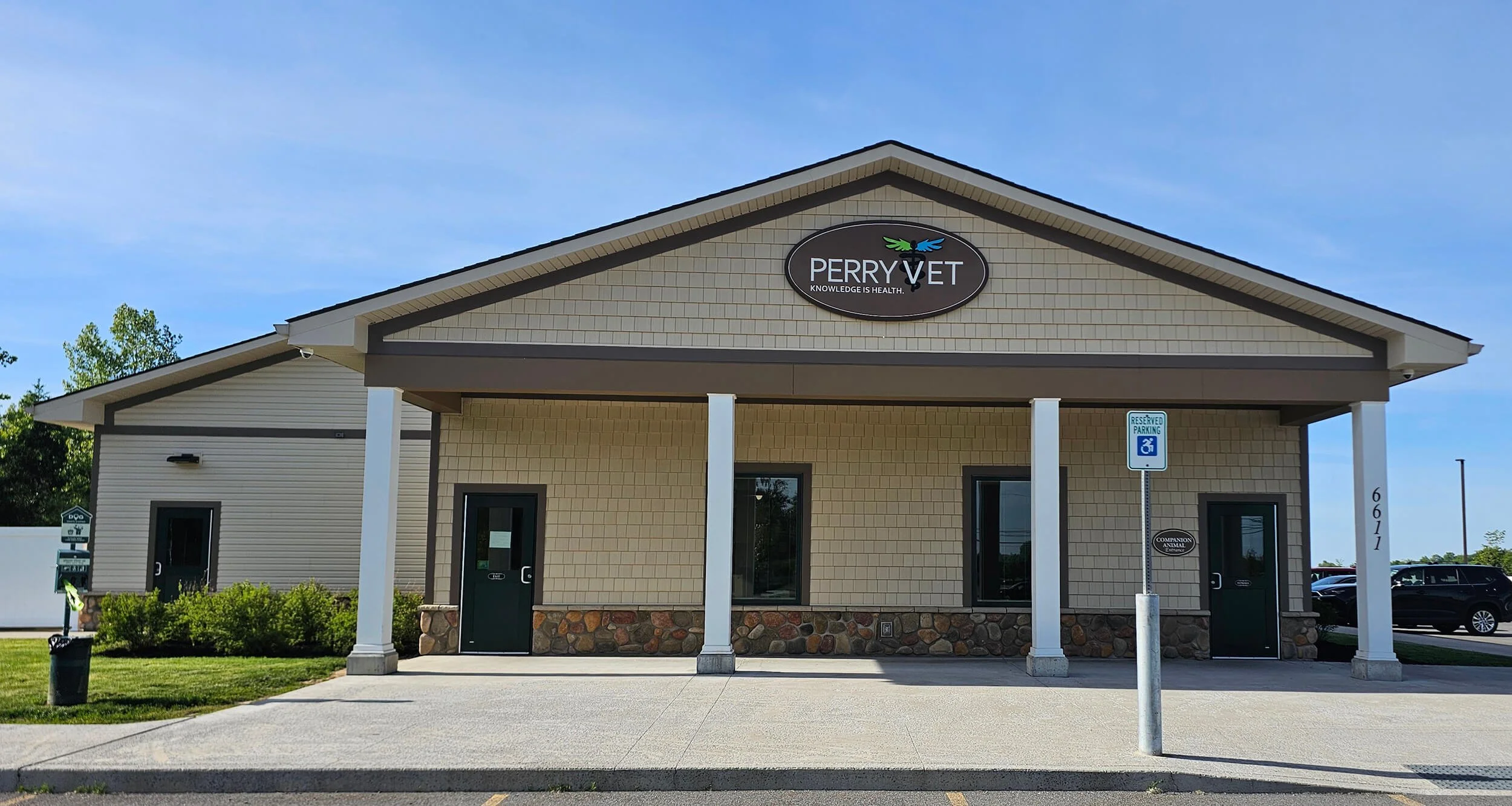 Front view of a veterinary office building with a sign that reads 'Perry Vet' and a slogan 'Knowledge is Health'. There is a reserved parking sign with a wheelchair symbol in front, and the building has two green doors, one on each side of the entrance. The sky is clear and blue.