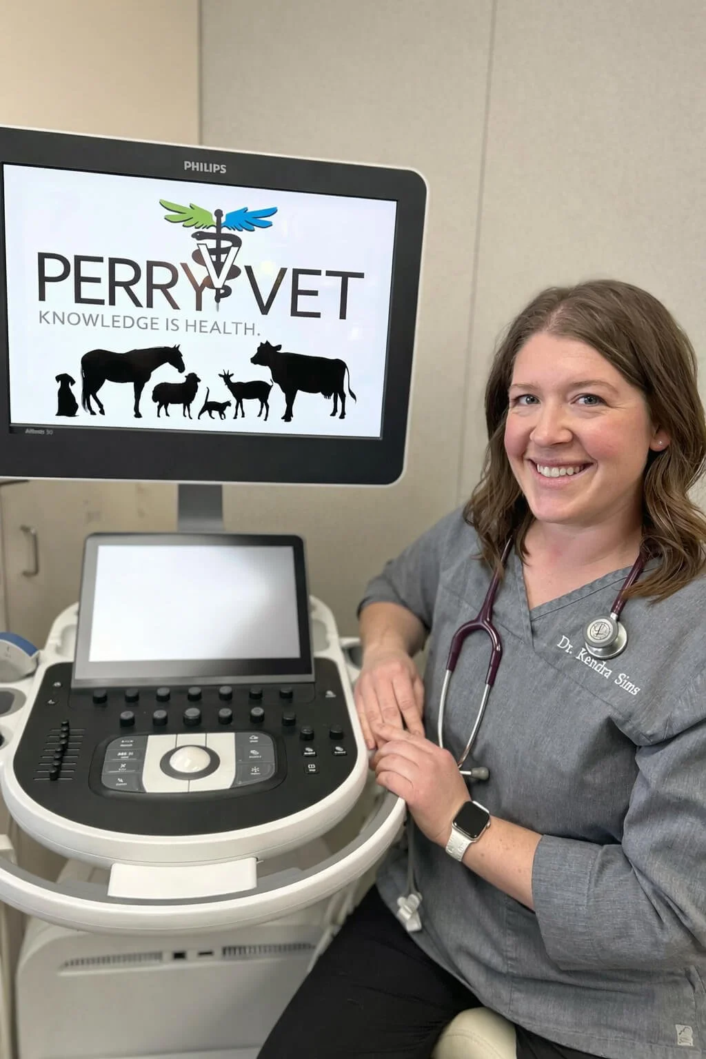 A smiling woman veterinarian with a stethoscope around her neck sitting next to an ultrasound machine at Perry Vet, with a screen displaying animals and the slogan "Knowledge is Health".