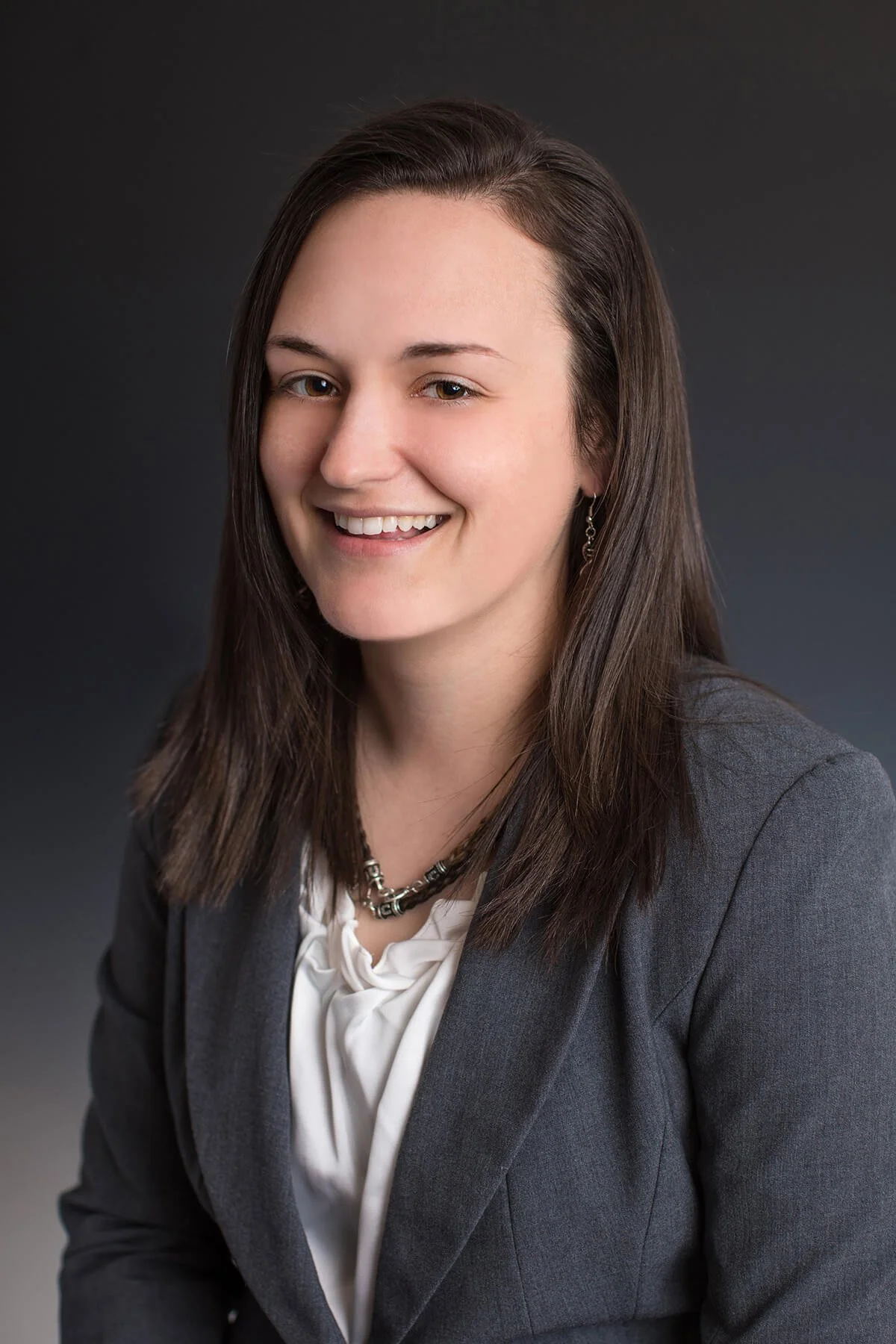 A young woman with long brown hair, smiling, dressed in a gray blazer over a white blouse, wearing a necklace and earrings, against a dark gray background.
