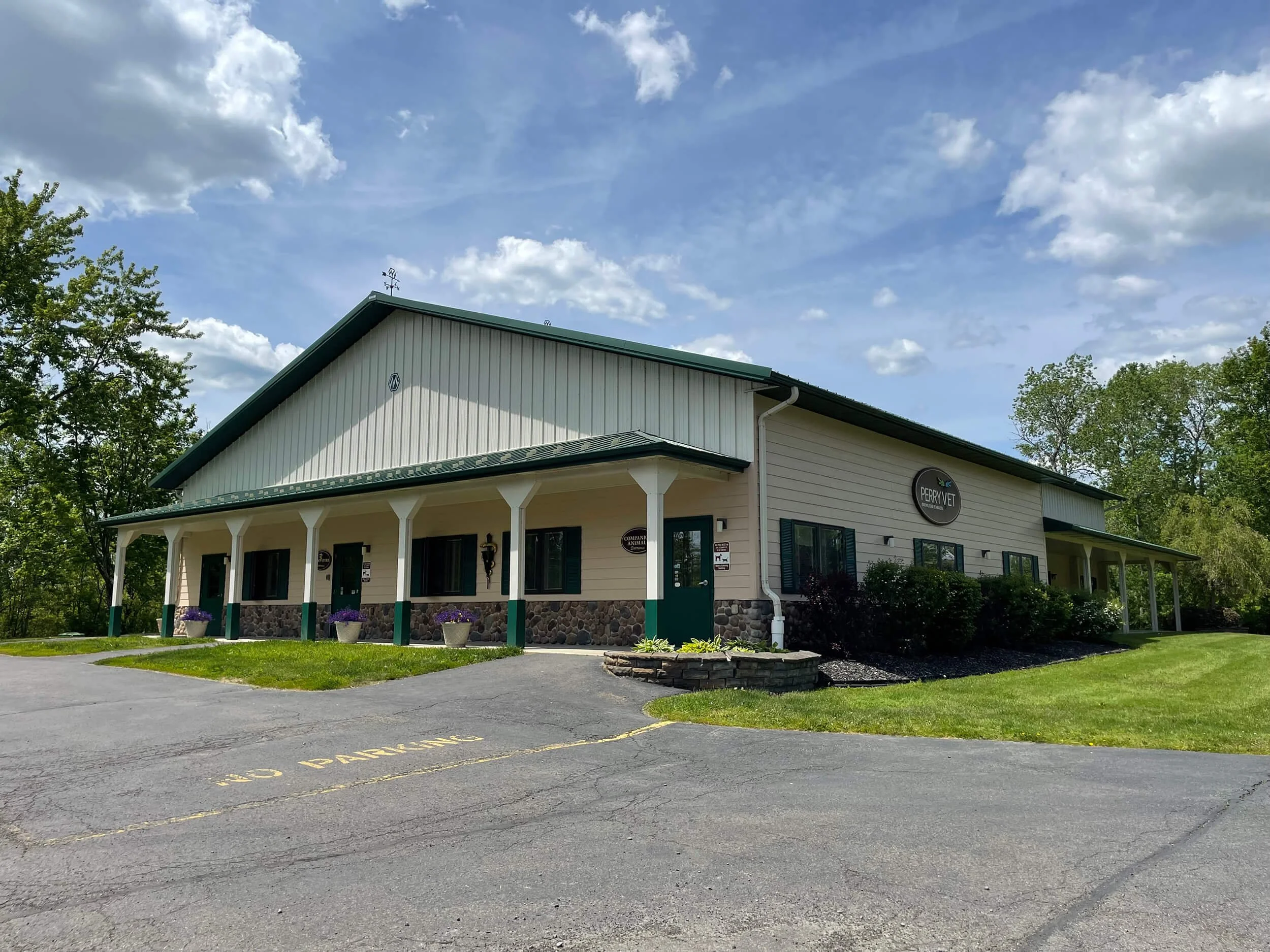 A two-story veterinary clinic building with a green roof and beige siding, surrounded by trees and a parking area with yellow painted markings.