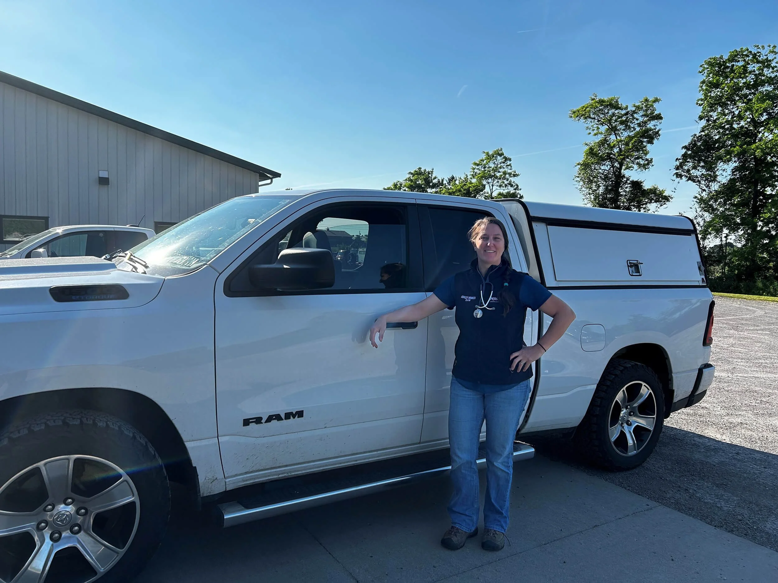 A woman standing next to a white RAM pickup truck in an outdoor parking lot, smiling at the camera on a sunny day with trees and a building in the background.