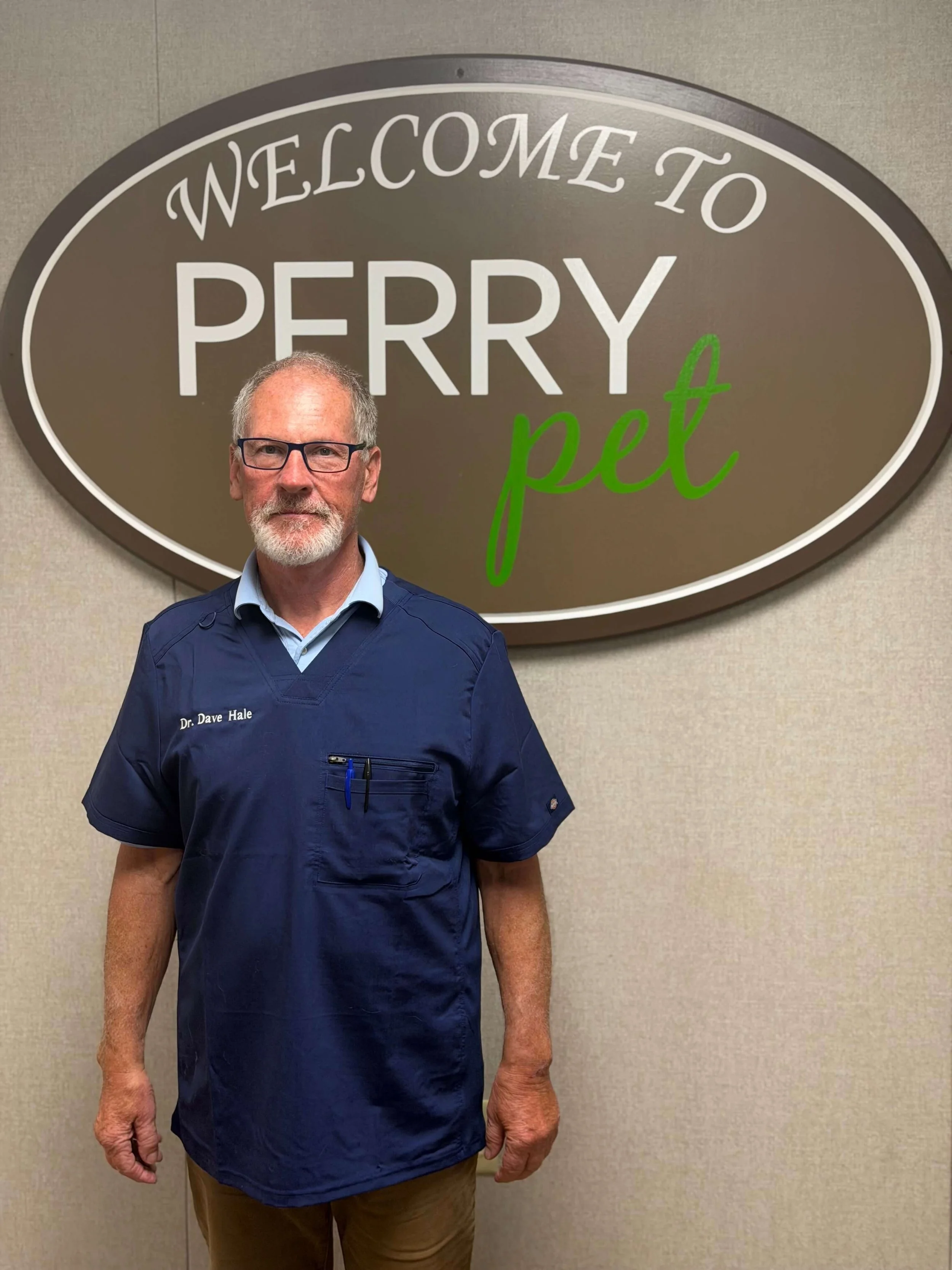 Older man in blue medical scrubs standing in front of a 'Welcome to Perry Pet' sign, indoors, beige wall background.