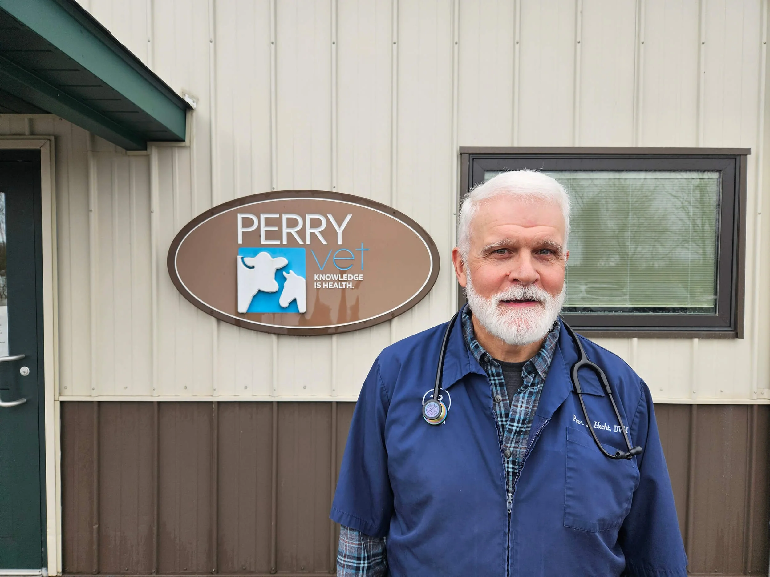 A man with white hair and a beard wearing blue medical scrubs, a stethoscope around his neck, standing outside a veterinary clinic. The clinic sign reads 'PERRY Vet' with a heart shape and animal silhouettes, and the slogan 'Knowledge is Health.'