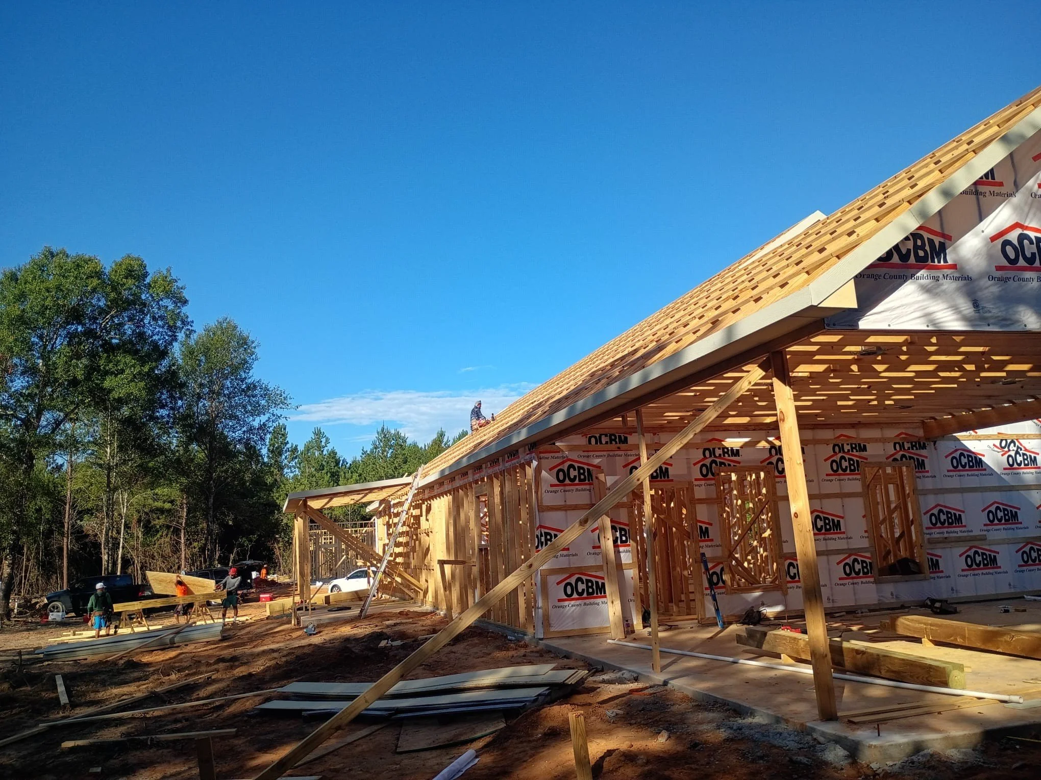 Construction site with a wooden framing structure in progress, workers on site, and trees in the background, under a clear blue sky.