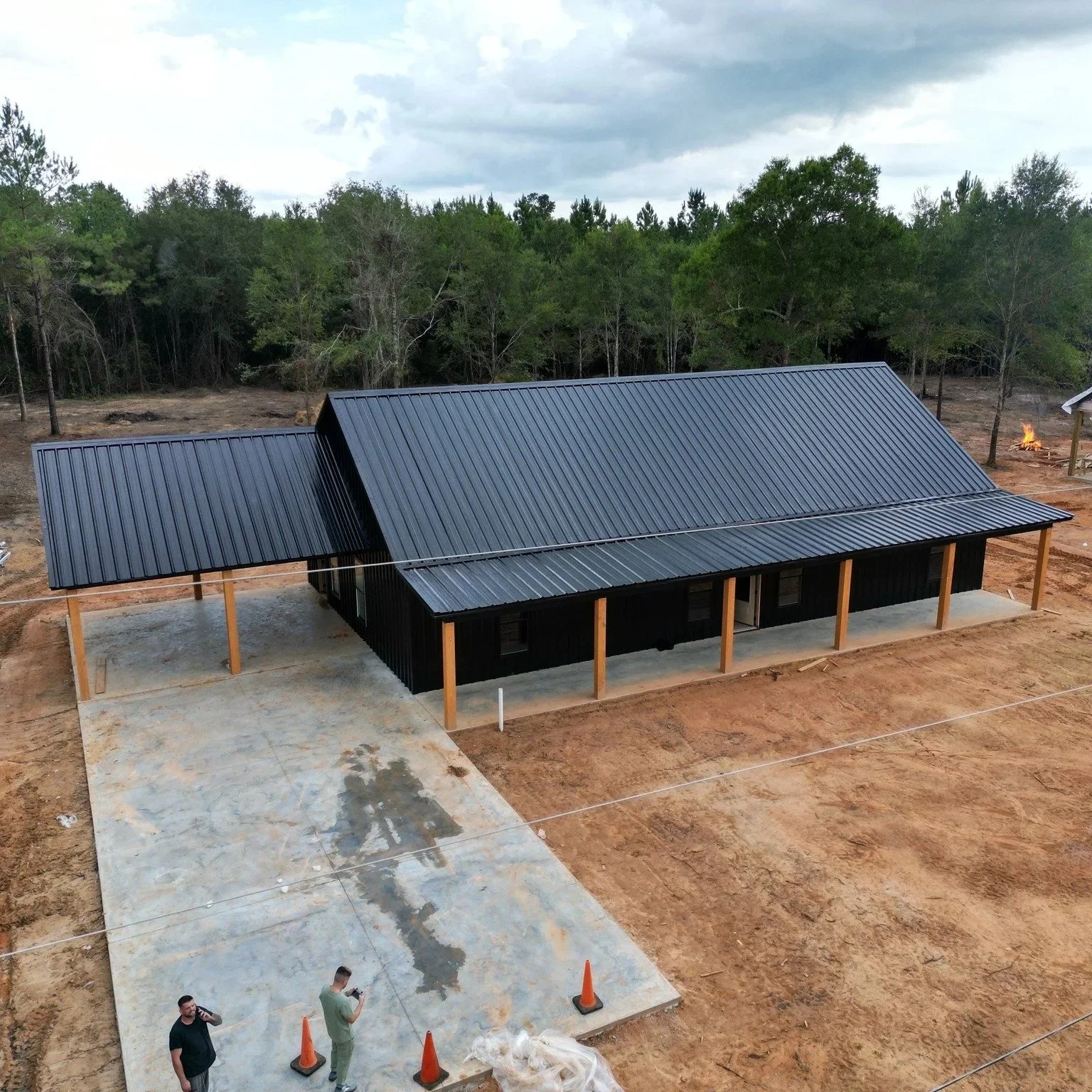 A house with black exterior walls and a metal roof under construction on an open lot, with construction workers, orange cones, and a concrete driveway in the foreground, and a wooded area in the background.