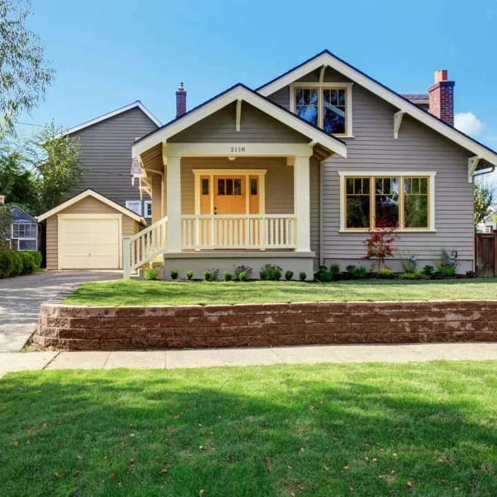 Front view of a gray, two-story house with a front porch, a small lawn, and a detached garage in the background.