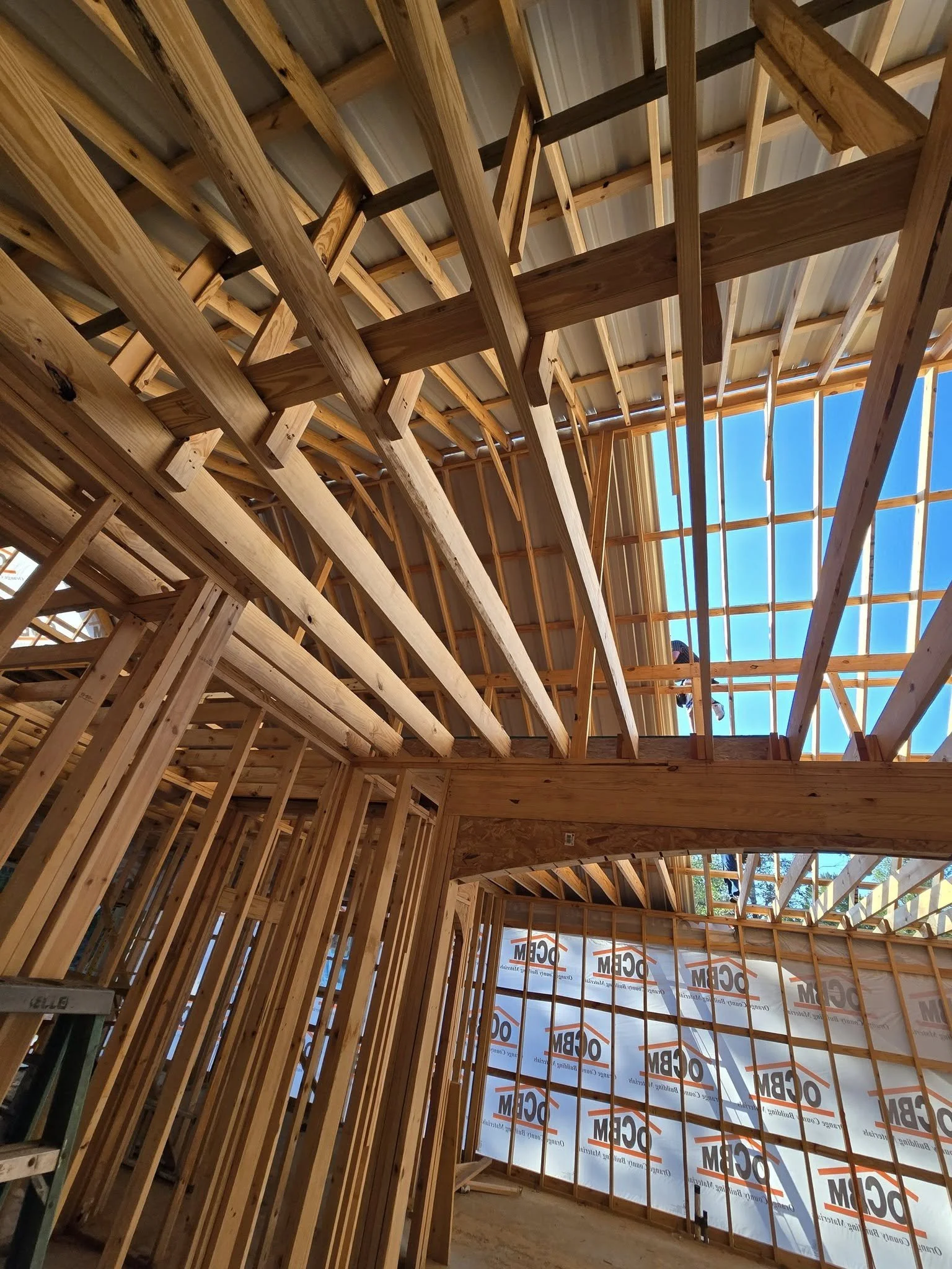 Interior view of a house under construction with exposed wooden framing and a partially completed roof with blue sky visible through the roof opening.