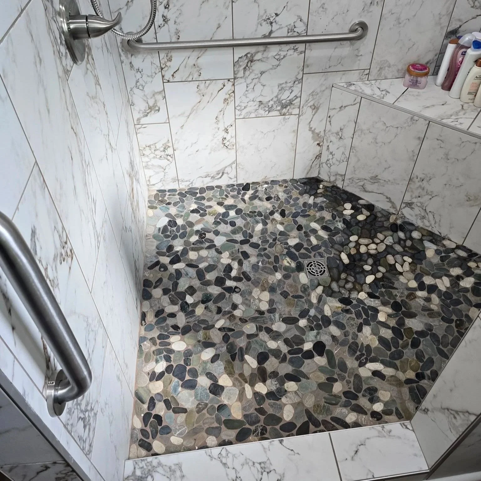A shower area with white marble tiles on the walls and a pebble stone floor, featuring a drain in the center. Contains a metal grab bar on the wall and toiletries on a built-in shelf.