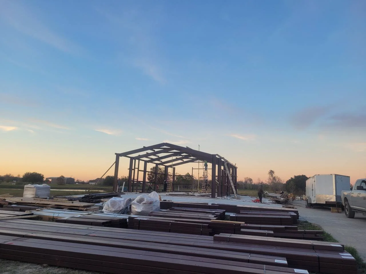 Construction site with a metal frame building under construction, building materials and lumber on the ground, parked vehicles, and a sunset sky.