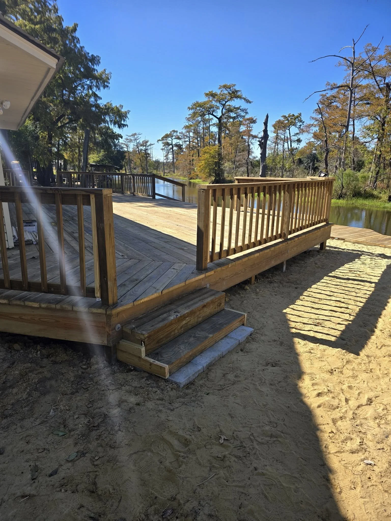 A wooden deck with steps leading down to sandy ground by a body of water, surrounded by trees under a clear blue sky.