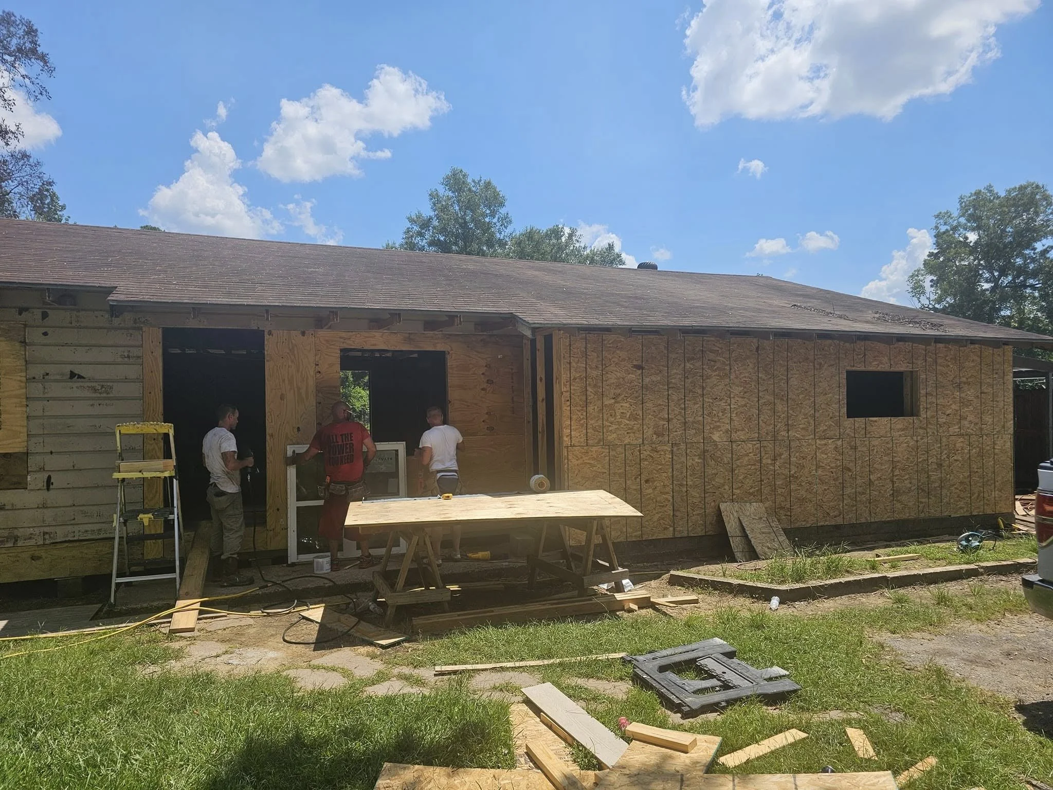 Construction workers building a house, with plywood sheathing on the exterior walls and construction tools and materials on the ground.
