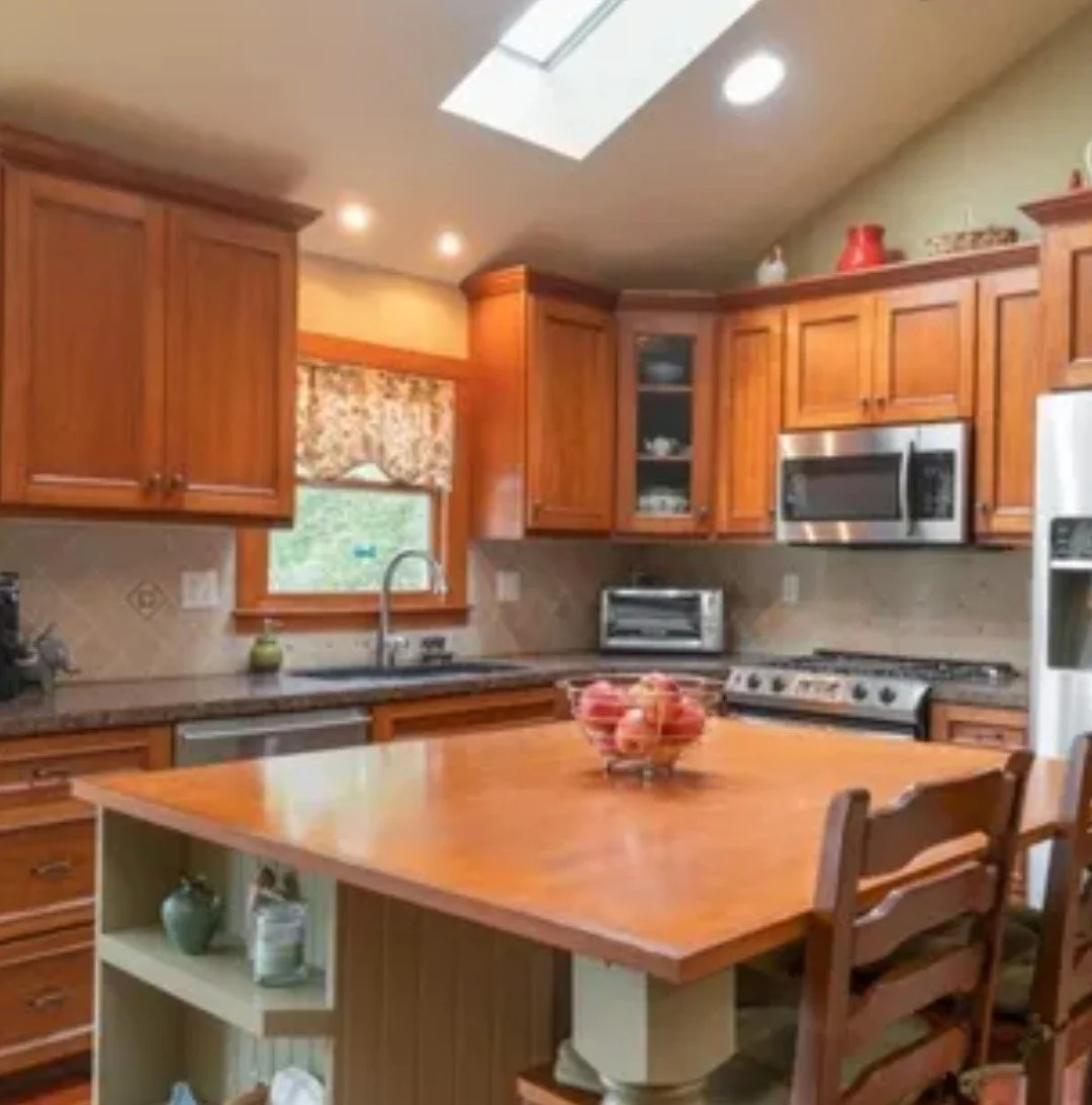 Kitchen with wooden cabinets, countertop, window with floral valance, stainless steel microwave, oven, and refrigerator, kitchen island with fruit bowl, wooden chairs.