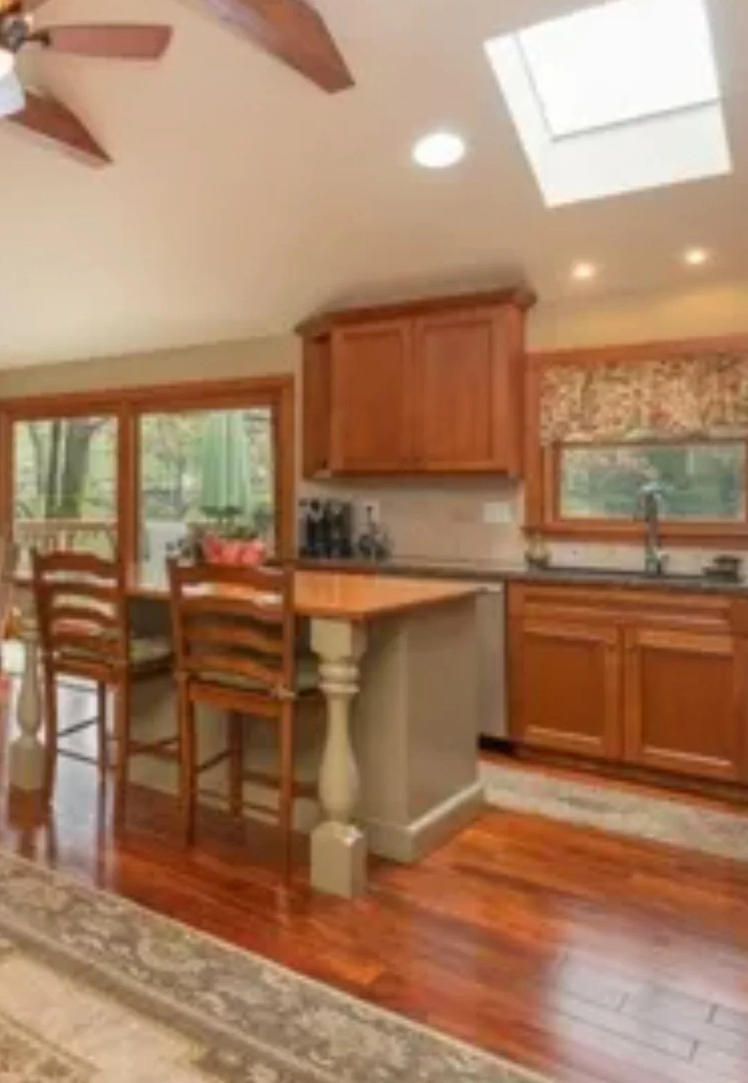 A cozy kitchen with wooden cabinets, a small island with bar stools, a sink under a window, and a sliding glass door leading to a deck with trees outside.
