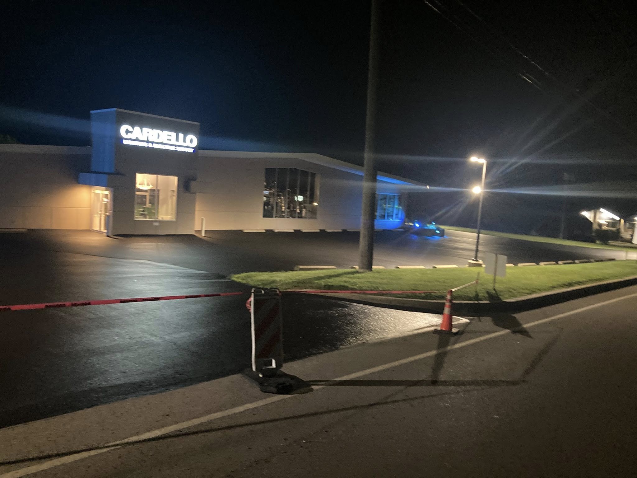 Nighttime scene of a commercial building with a lit sign reading 'CARDELLO' and a large glass window, with a few street lights and orange cones blocking part of the parking lot.