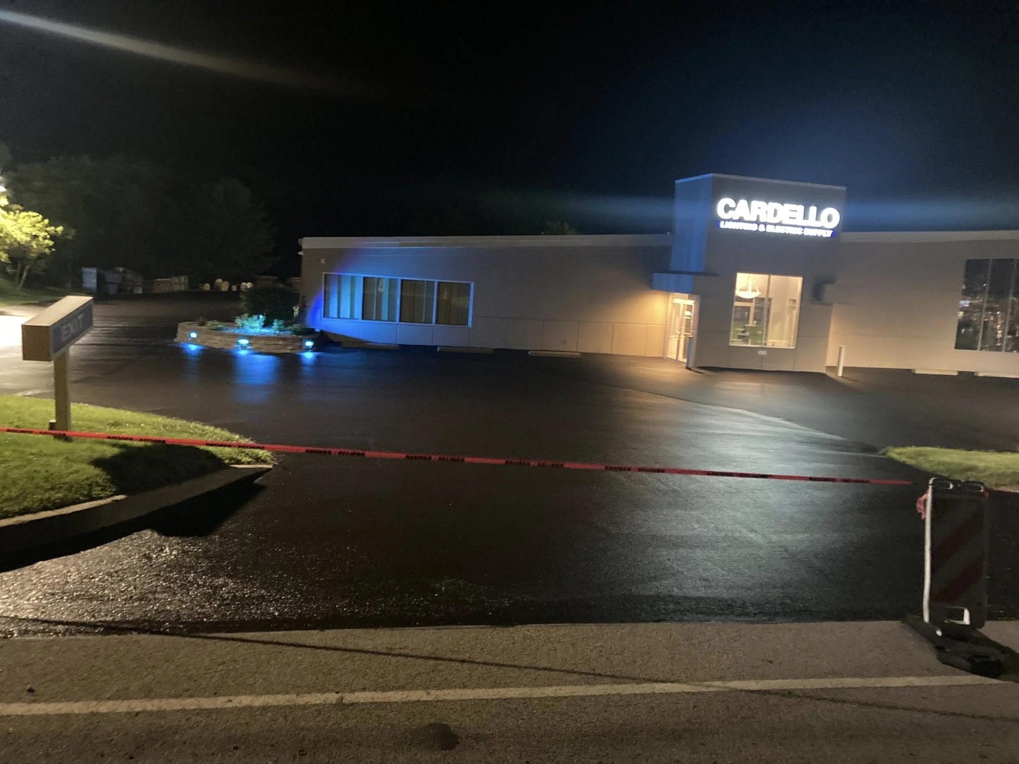 Nighttime exterior shot of a building with a lit sign reading 'CARDELLLO,' featuring blue and yellow lights illuminating nearby plants, with a wet parking lot and a red and white barricade across the entrance.