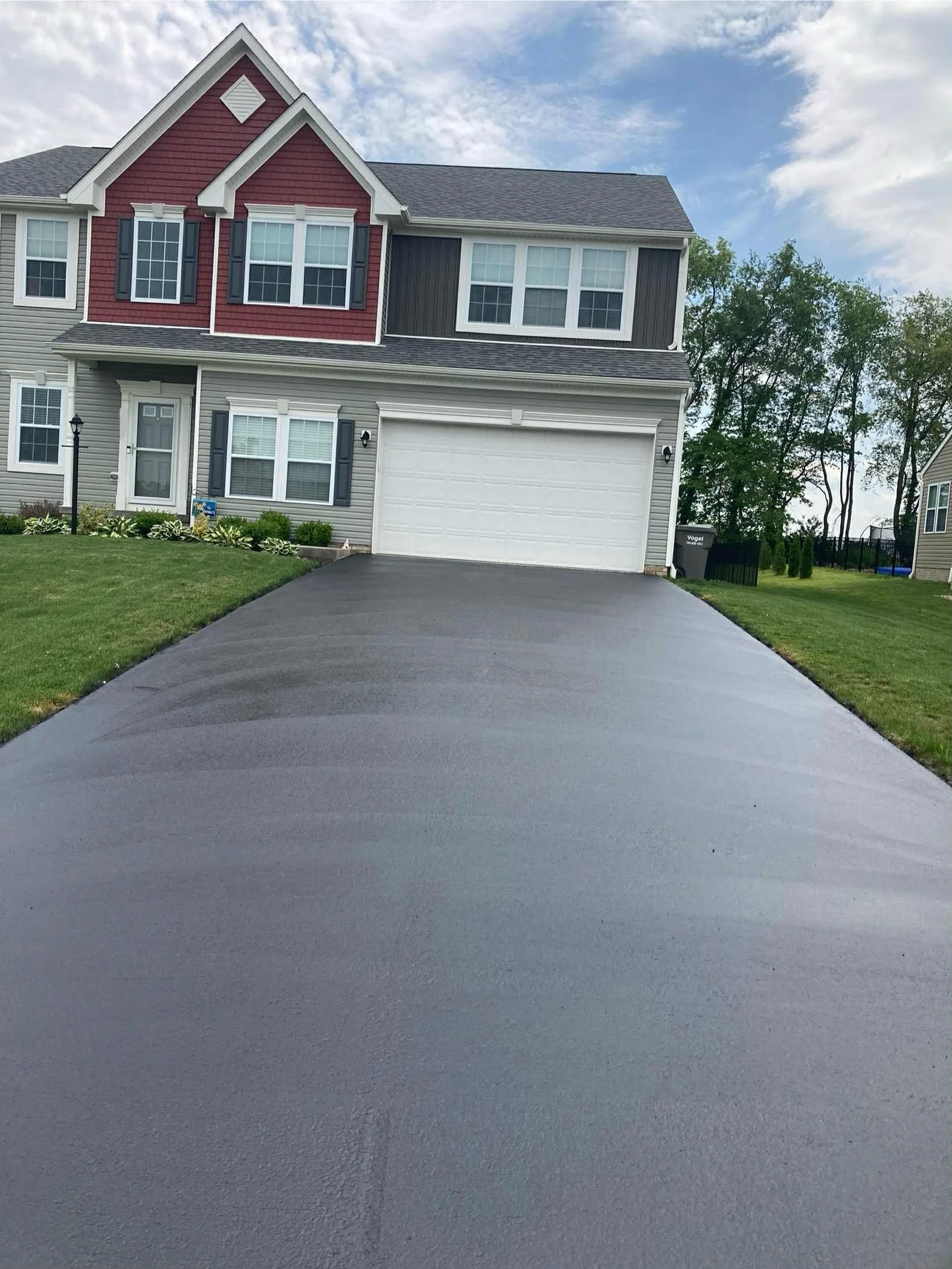 Photo of a newly paved blacktop driveway leading up to a two-story residential house with gray and red siding, white garage door, and windows. The house has black shutters and is surrounded by green grass and small plants.