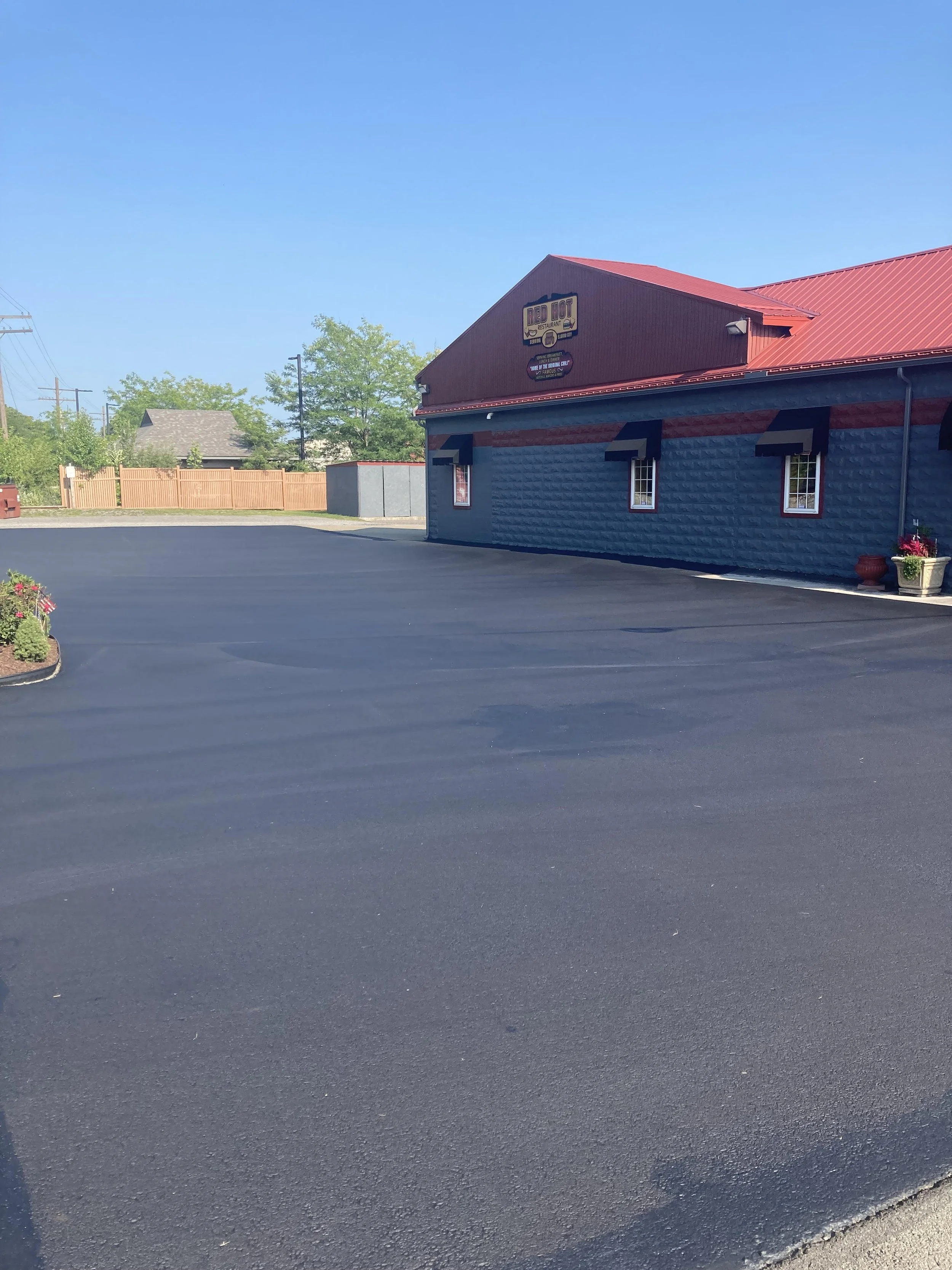 A paved parking lot outside a restaurant called Red Hot. The restaurant building has a red roof and blue siding with red accents, and there are plants near the building. Clear blue sky and trees are visible in the background.