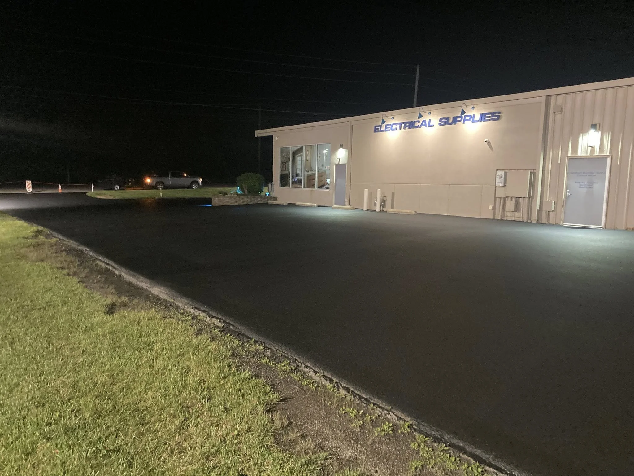 Night view of a building labeled "Electrical Supplies" with a lit parking lot and some parked cars in the background.