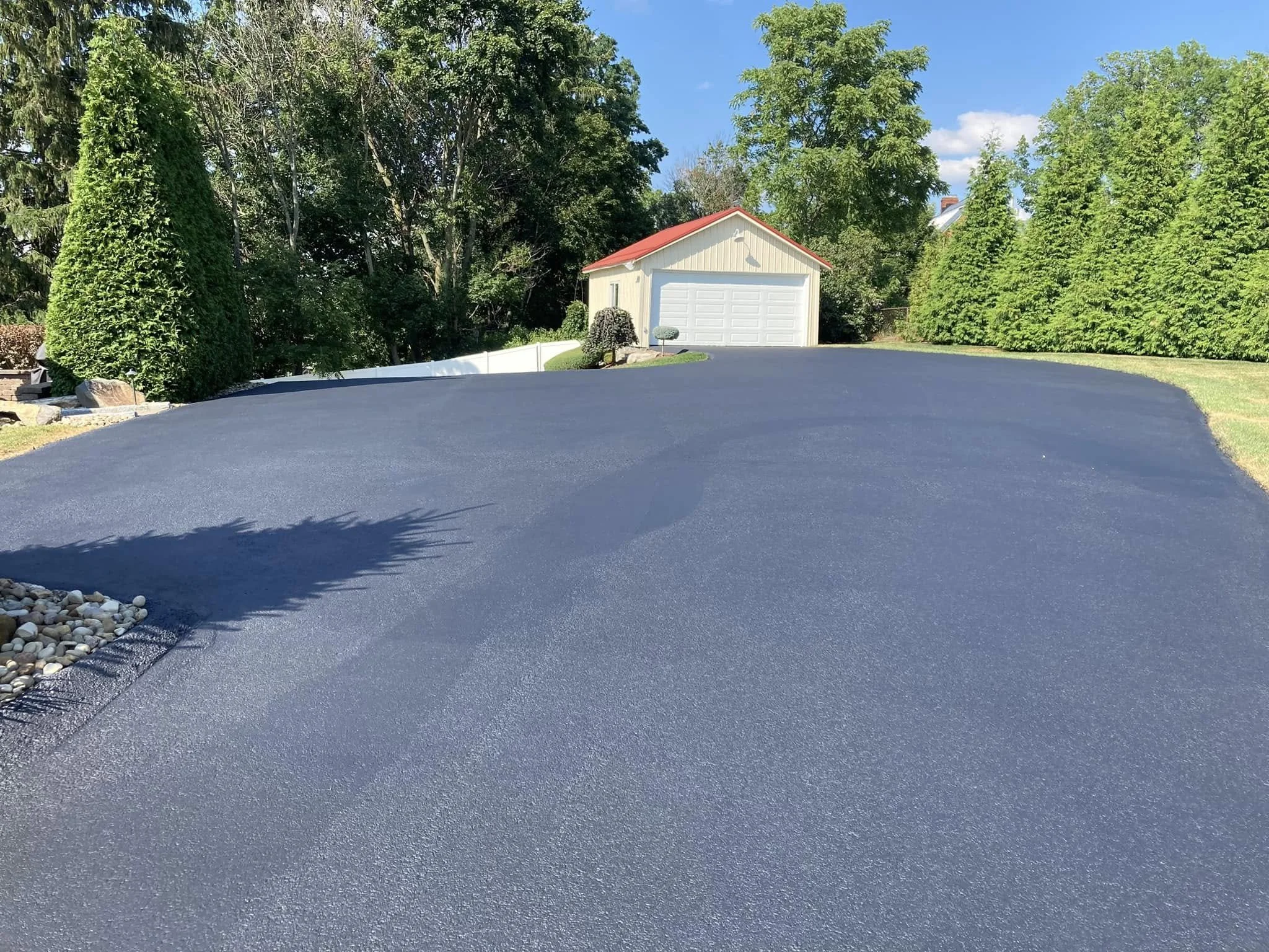 A freshly paved black asphalt driveway leading to a white garage with a red roof, surrounded by green trees and grass on a clear day.