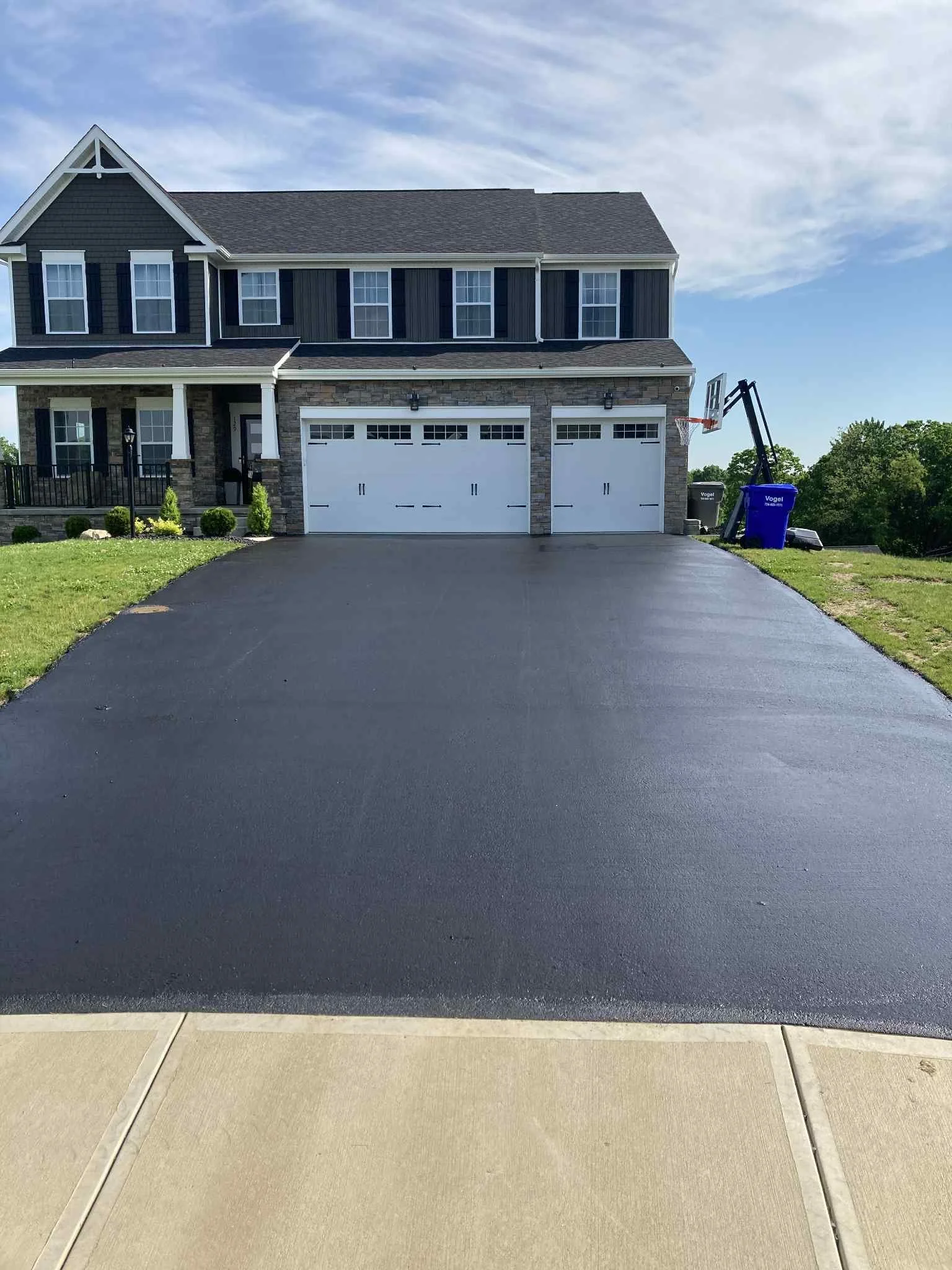 Freshly paved black asphalt driveway in front of a large two-story house with a three-car garage, front porch, and a basketball hoop on the right side near a blue trash bin, under a partly cloudy blue sky.