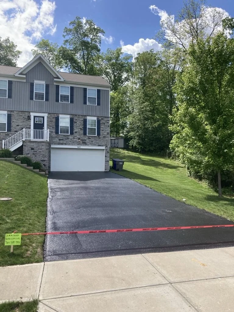Newly paved driveway in front of a two-story gray house with white and black shutters, surrounded by green grass and trees, with caution tape across the sidewalk.