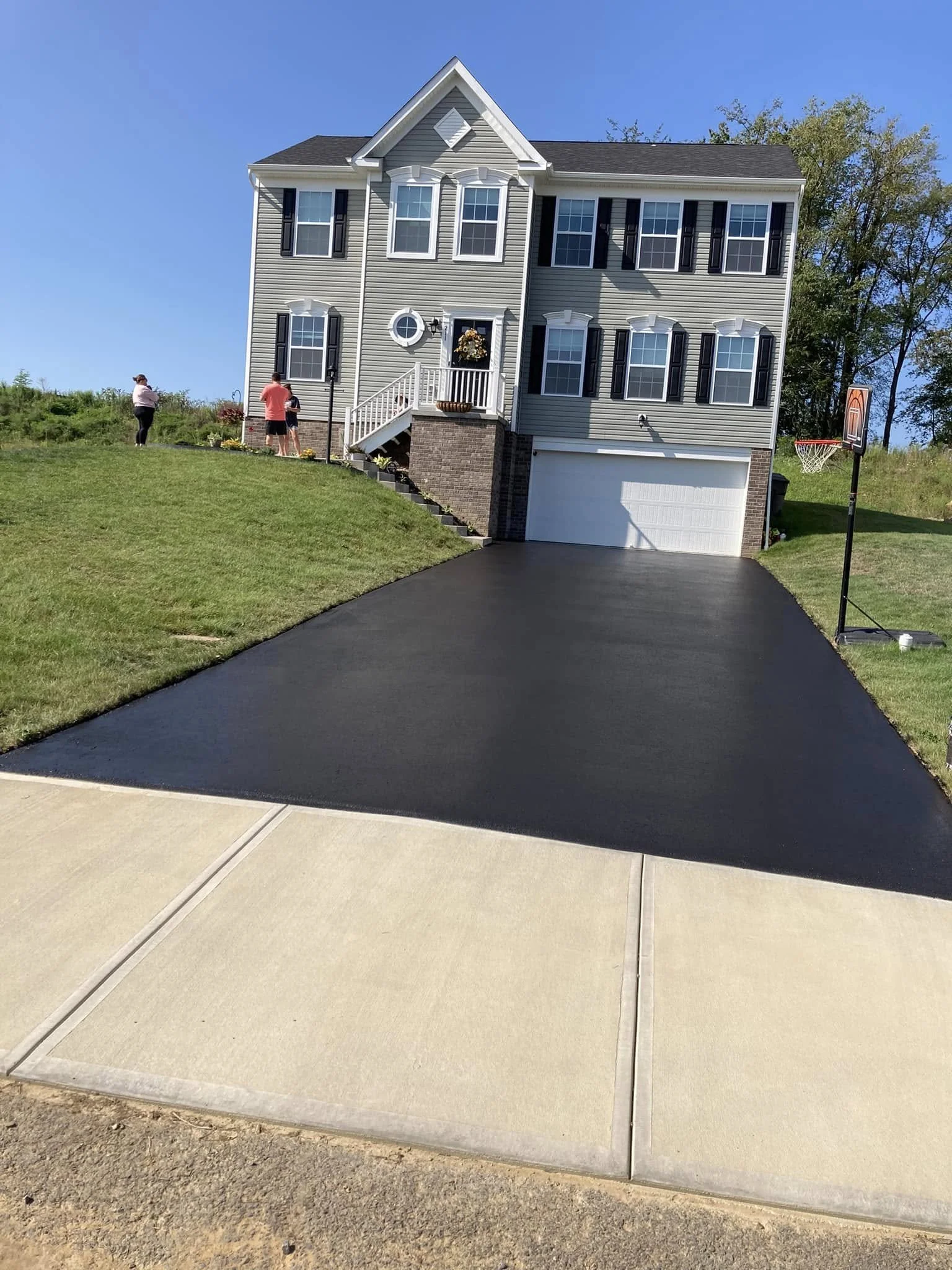 A newly paved black asphalt driveway slopes up to a two-story house with gray vinyl siding, white trim, and black shutters. The house has a small front porch and a garage. To the right, a portable basketball hoop is set up on the lawn. The sky is cle