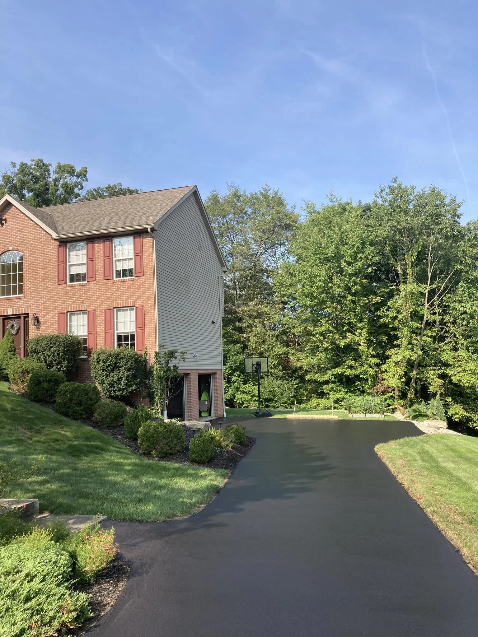 A newly paved black asphalt driveway leading up to a brick and siding house with red shutters, surrounded by green bushes and tall trees, under a clear blue sky.