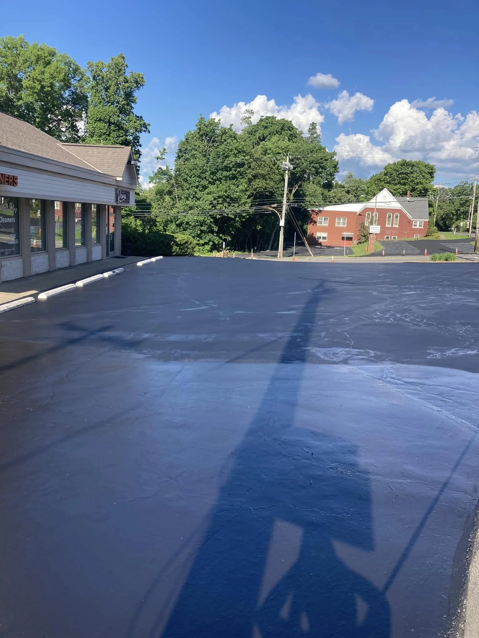 Empty parking lot with fresh black asphalt, adjacent to a building with large windows, under a bright blue sky with white clouds, surrounded by green trees and some brick buildings in the background.