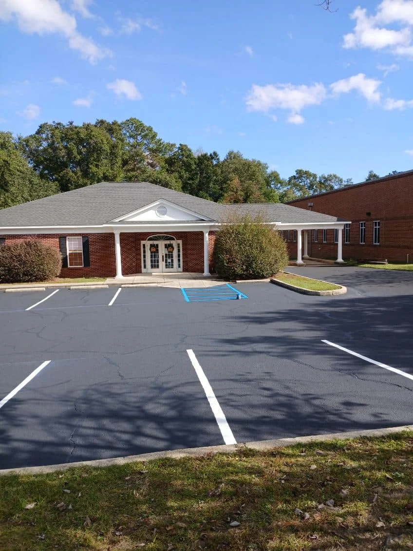Empty parking lot with three empty spaces and a handicapped spot, in front of a brick building with white columns and a gray roof, under a blue sky with scattered clouds.