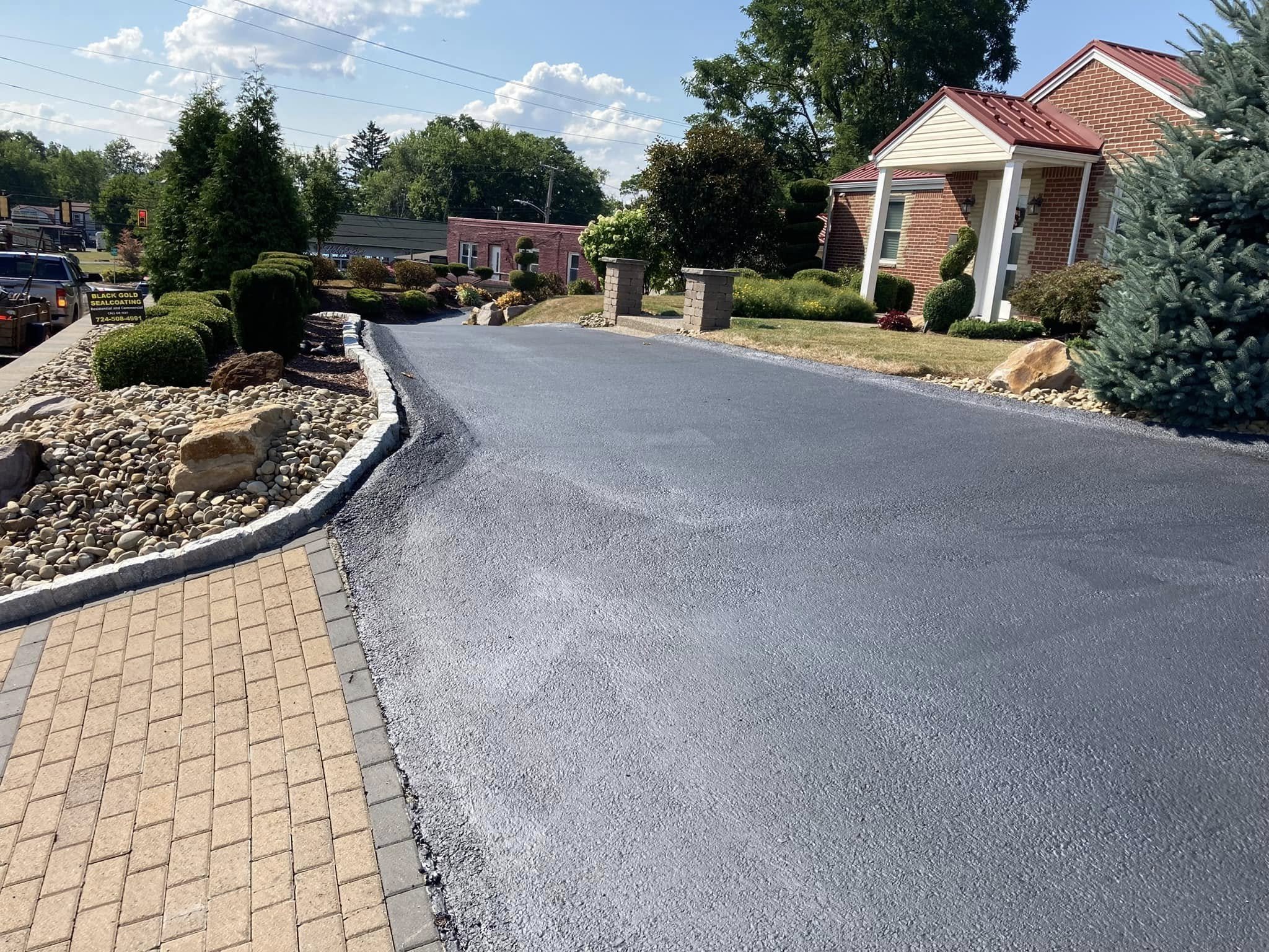A freshly paved black asphalt driveway in front of a brick house with a red metal roof, surrounded by landscaped bushes, trees, and decorative rocks on a sunny day.