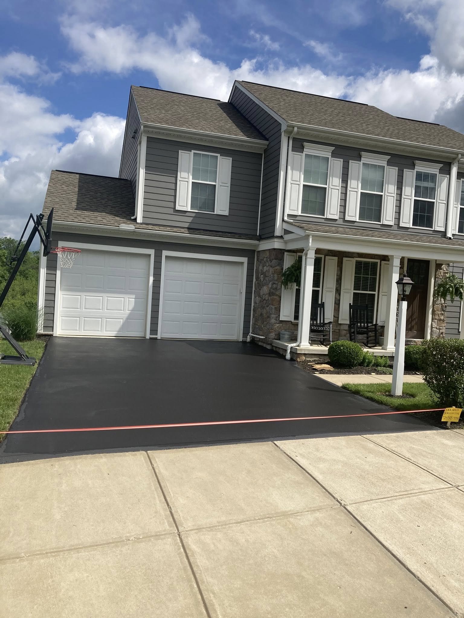 Newly paved black driveway in front of a gray two-story house with white trim, stone facade, and a small front porch, under a partly cloudy sky.