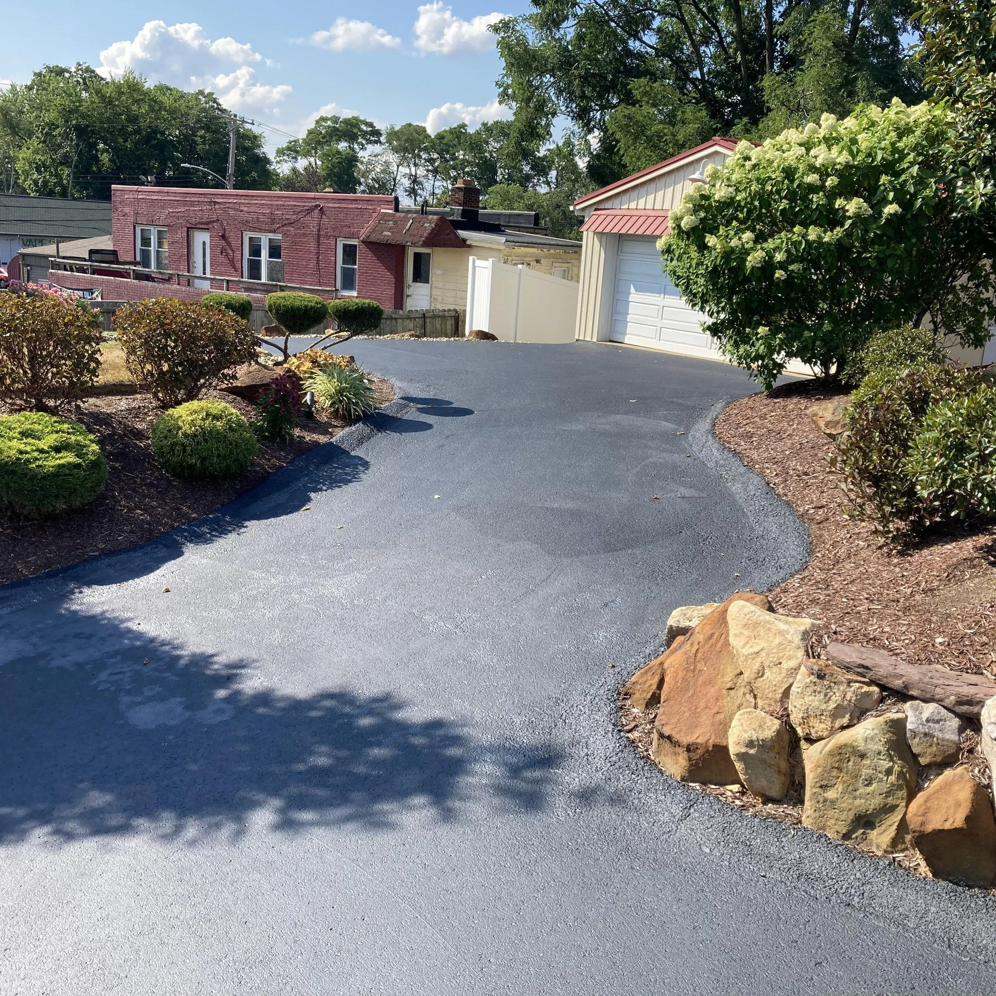 Paved driveway with landscaped plants and rocks, leading to a garage and surrounding houses on a sunny day.