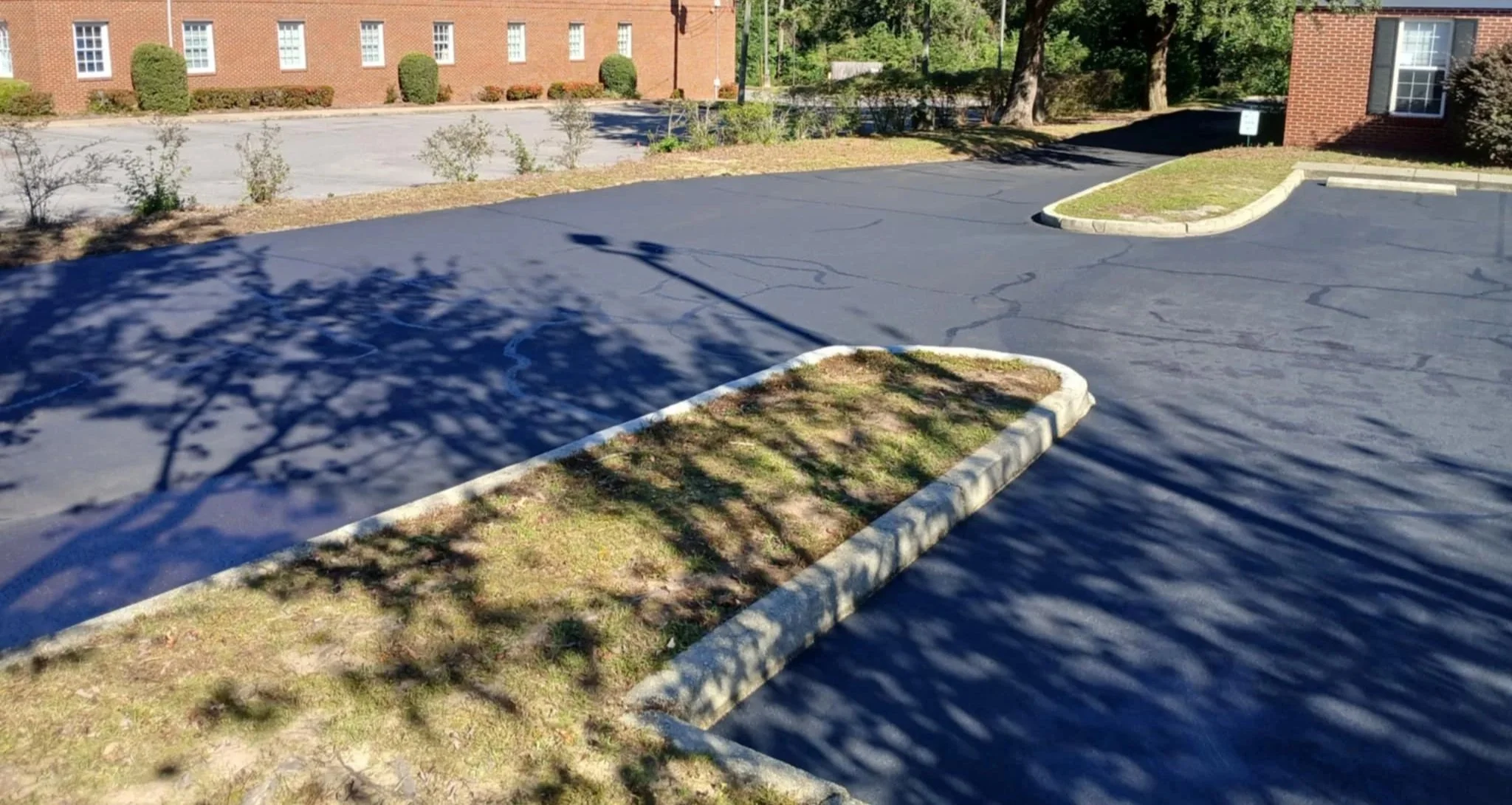 Empty parking lot with small landscaped islands, brick building in background, and shadows from trees and a streetlamp on the asphalt.