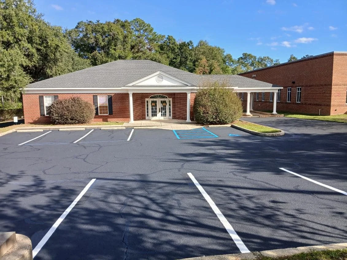 A brick building with white columns and multiple windows, surrounded by a parking lot with white lines designating parking spaces and a designated handicapped parking spot marked with blue paint.