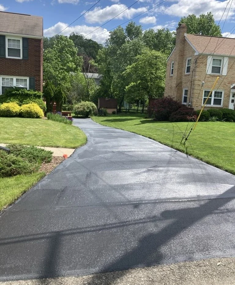 Newly paved black asphalt driveway in front of brick and beige houses, with green lawns and trees on either side, under a partly cloudy sky.