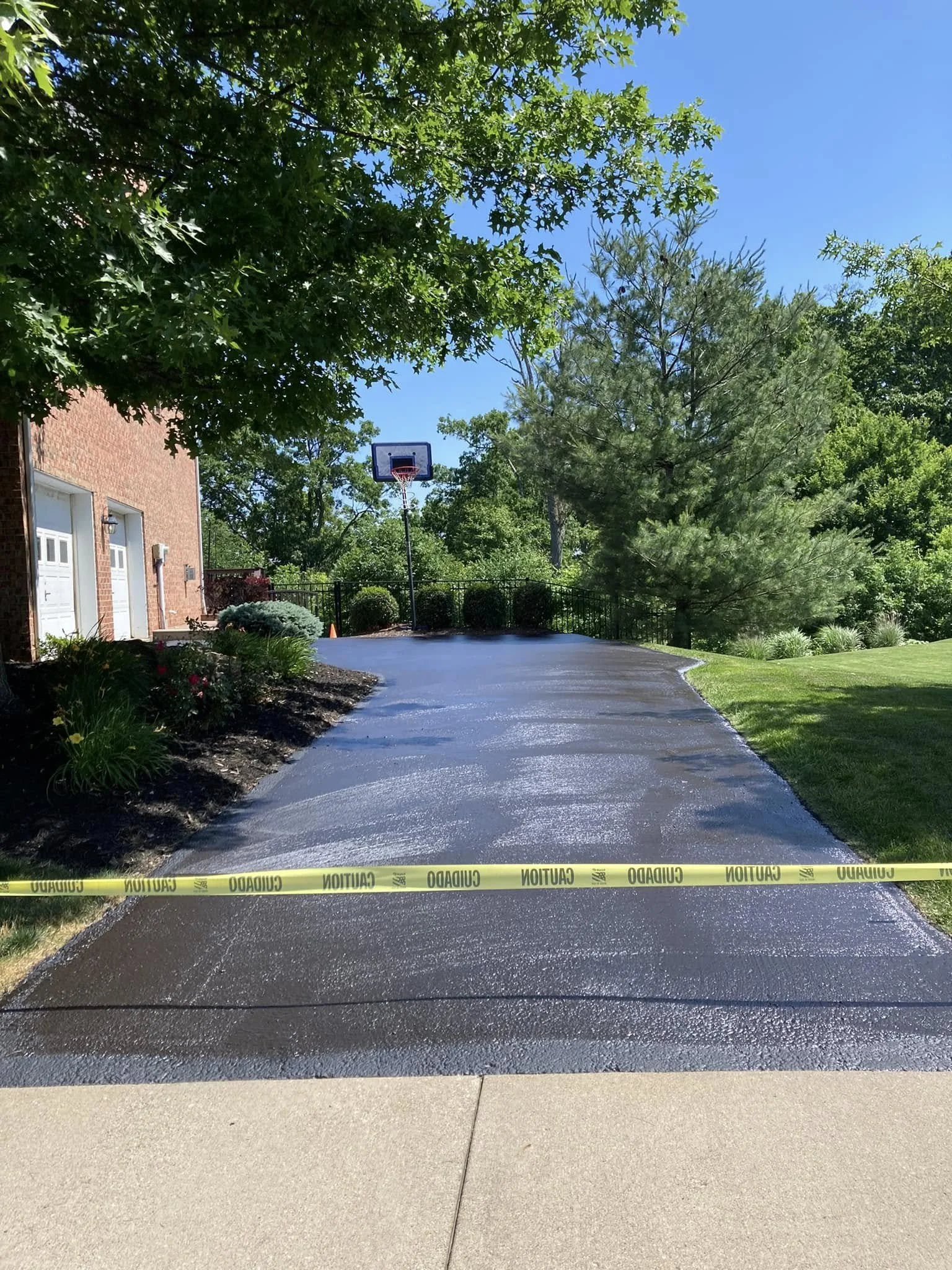 Freshly paved driveway with caution tape across the front, a basketball hoop at the end, surrounded by greenery and trees under a blue sky.