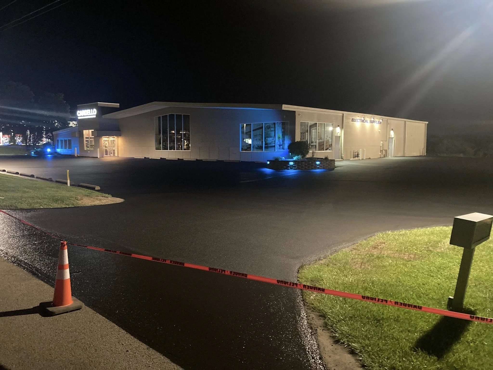 Photo of a modern commercial building at night, illuminated by exterior lighting, with a parking lot in the foreground. There is caution tape and an orange traffic cone near the edge of the parking lot.
