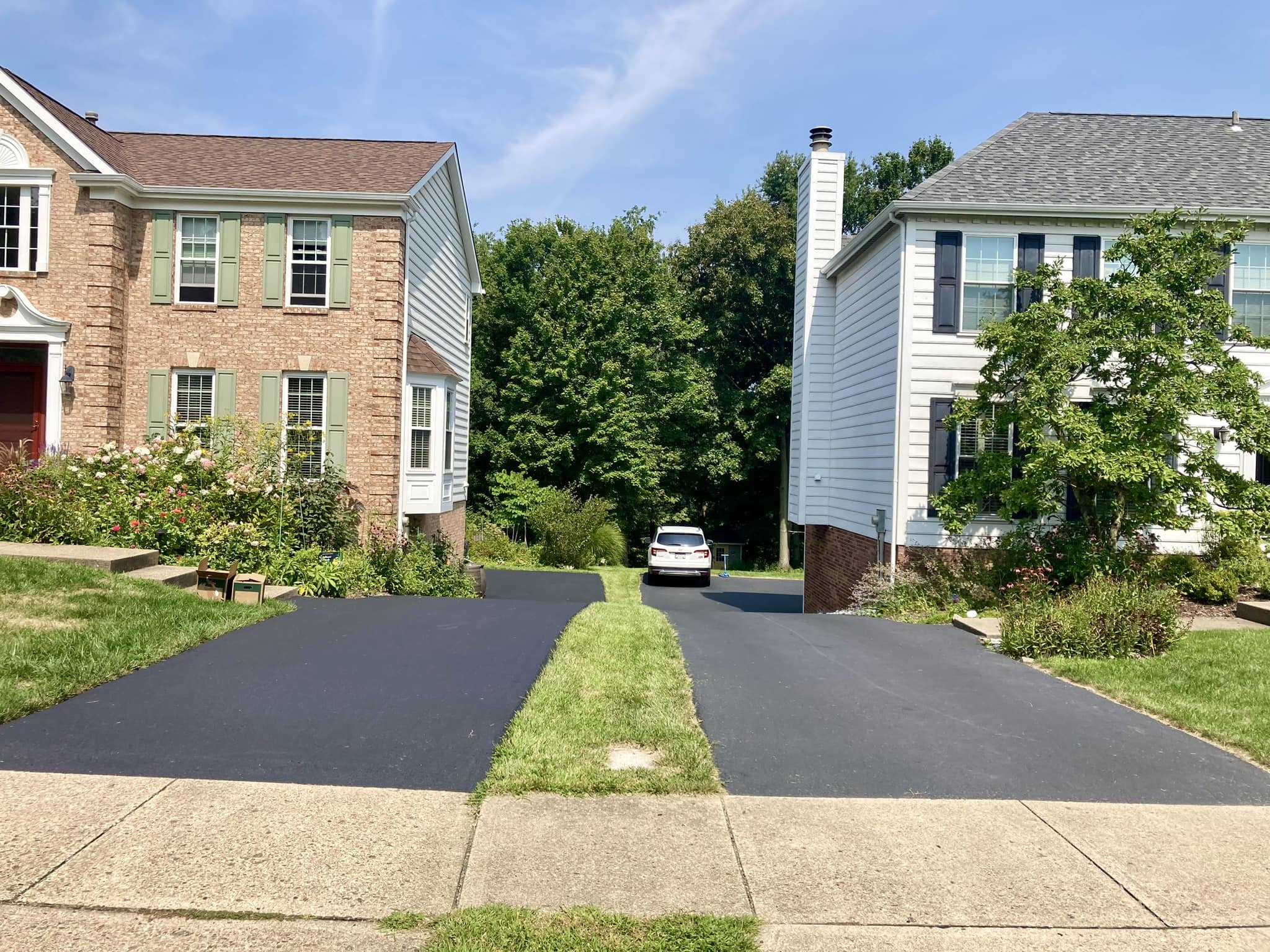 Two houses with driveways, a white car parked in the distance, green trees, and a partly cloudy sky.