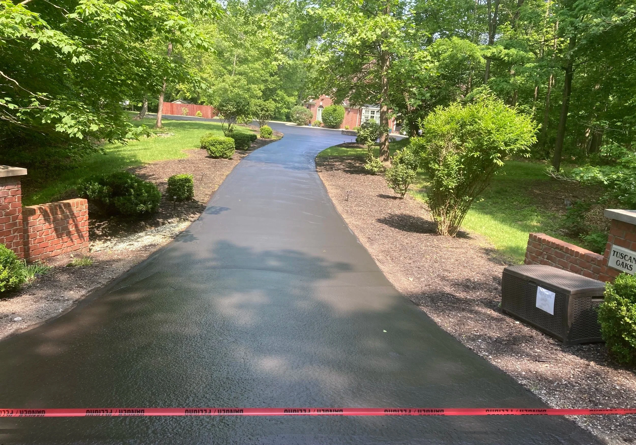 Paved black asphalt driveway in a residential neighborhood surrounded by green trees and shrubs, with brick walls on either side.